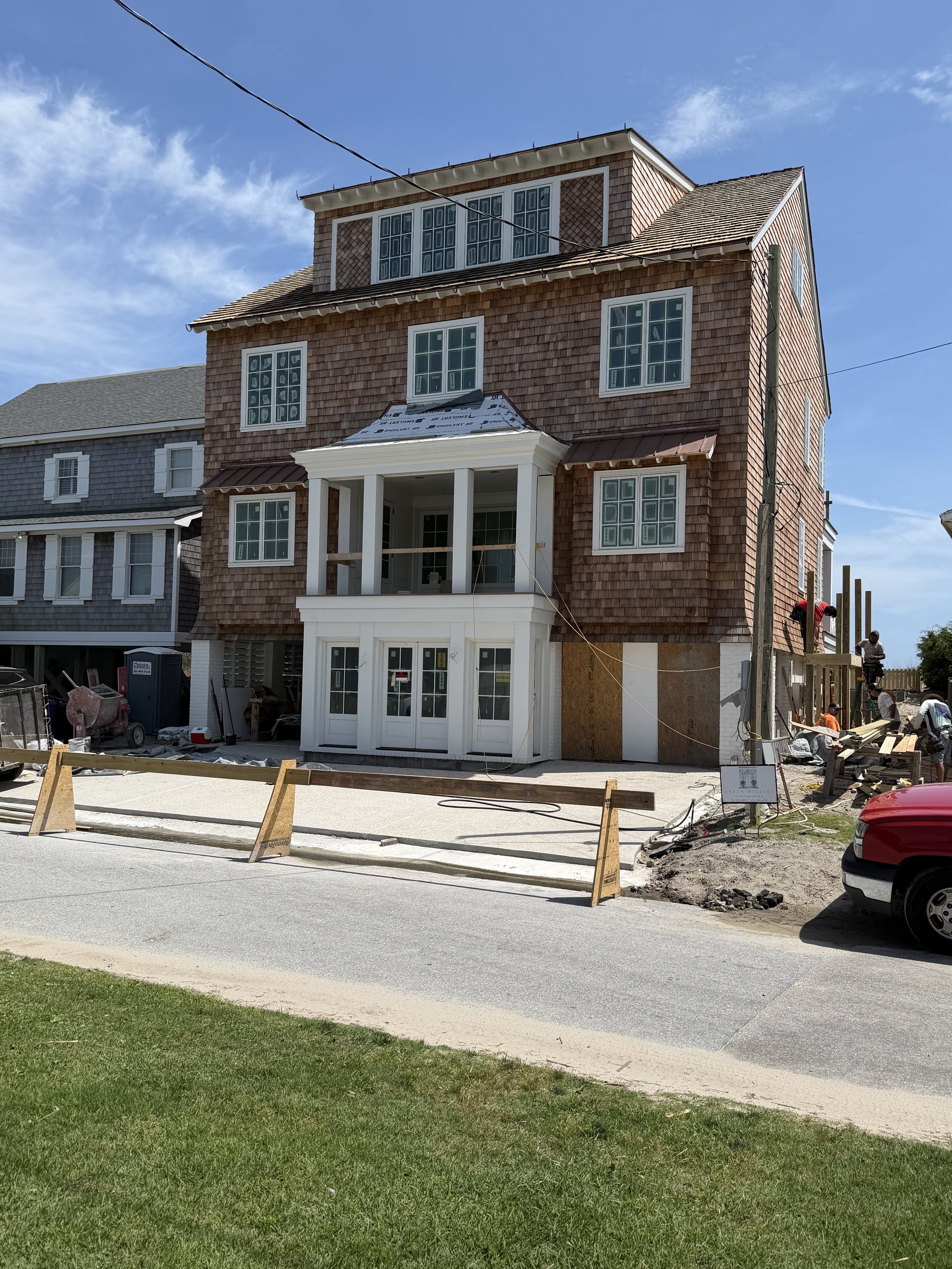 A multi-story house under construction with workers visible on the right side, surrounded by a wooden fence, with a driveway in front and a green lawn.