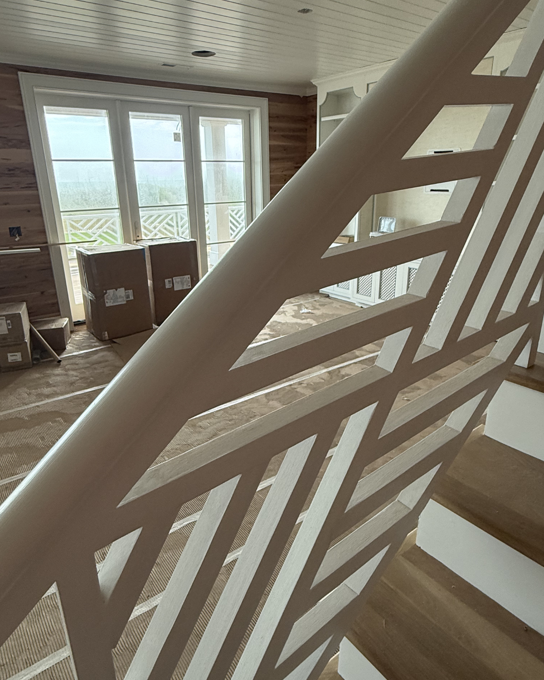 Interior of a house under construction with a staircase, cardboard boxes near large windows, and white cabinets in the background.