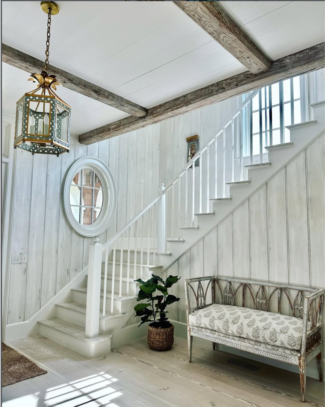 A bright entryway with white wooden paneled walls, a white staircase with wooden steps, and a large window letting in natural light. There is a vintage-style bench with a patterned cushion, a potted plant, a round mirror, and a decorative hanging lantern.