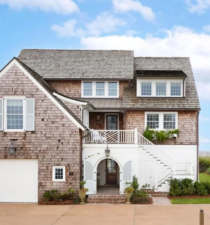 A two-story house with a shingle exterior, white trim, and multiple windows, including a prominent dormer, with stairs leading up to a covered front door, surrounded by potted plants and greenery.