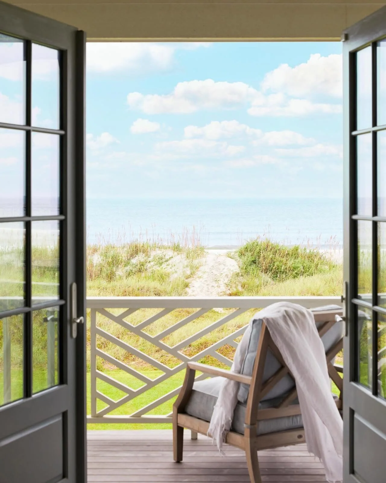 Open patio door looking out to a beach with sand dunes, grasses, blue sky, and clouds.