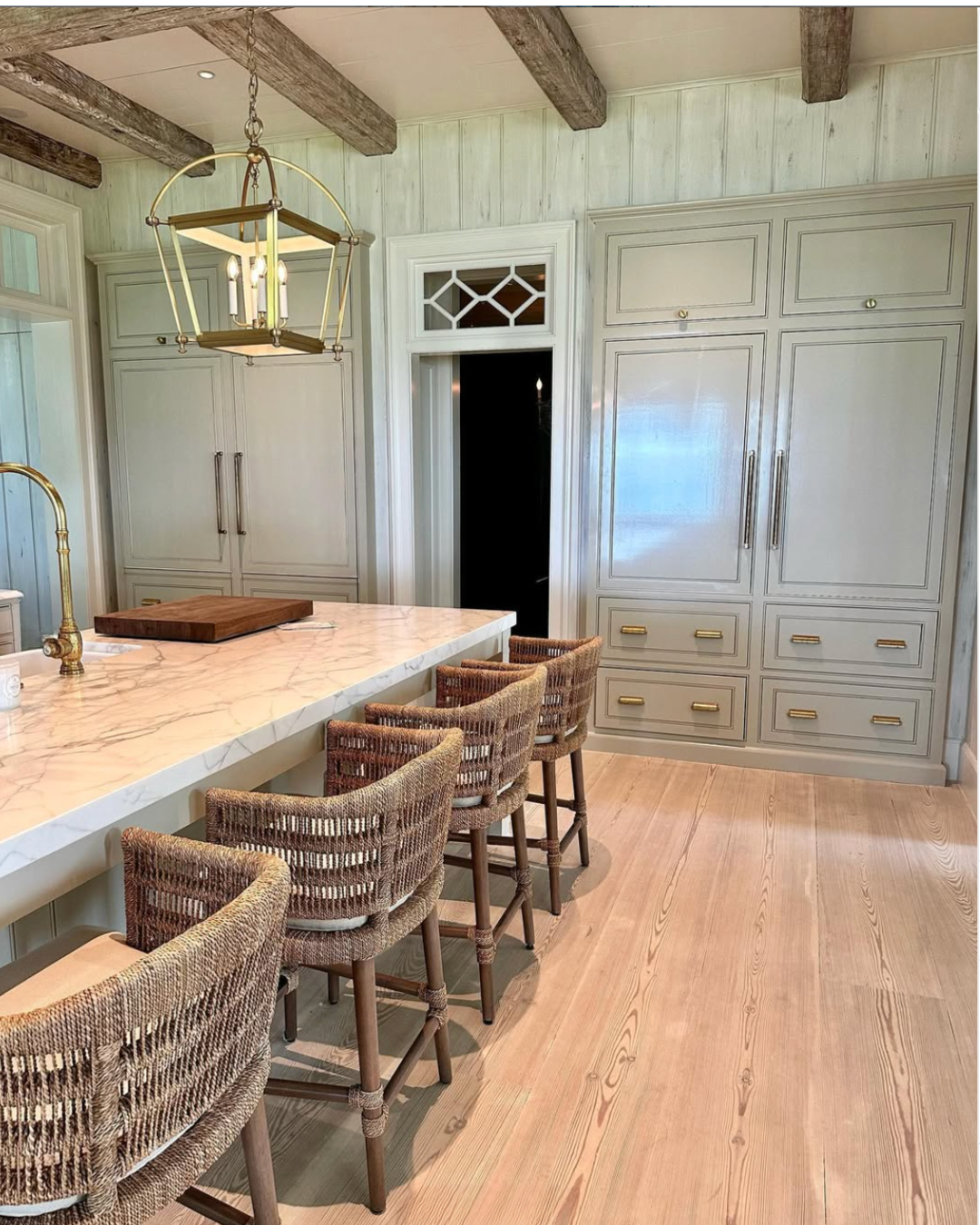 Kitchen with light gray cabinetry, wooden beams on the ceiling, a white marble island with a gold faucet, wicker bar stools, and a black doorway.