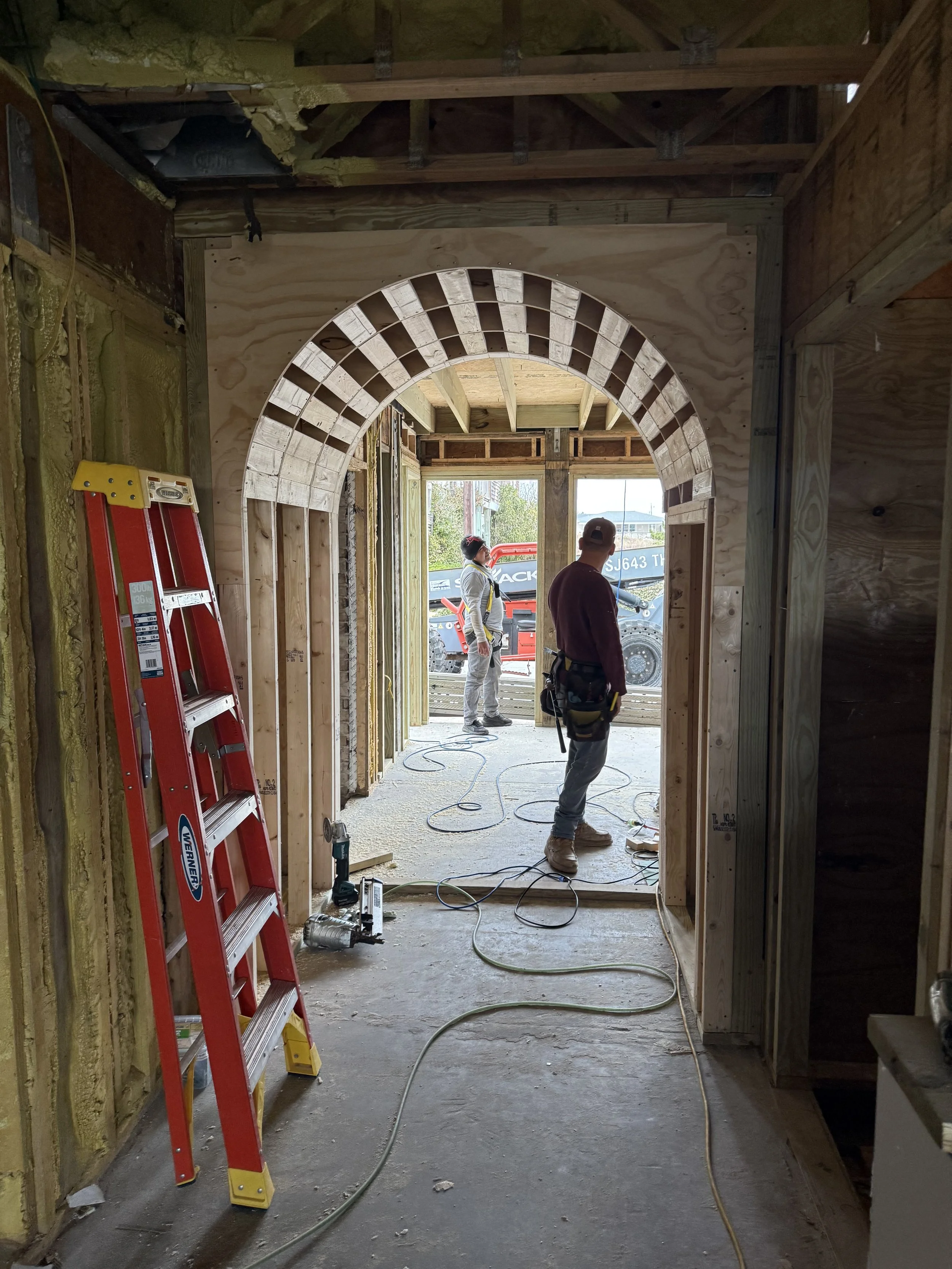 Inside a house under construction with wooden framing and an archway. Two workers are visible, with a ladder leaning against the wall on the left and construction tools on the floor. Outside, construction equipment and a boat trailer are seen through the opening.