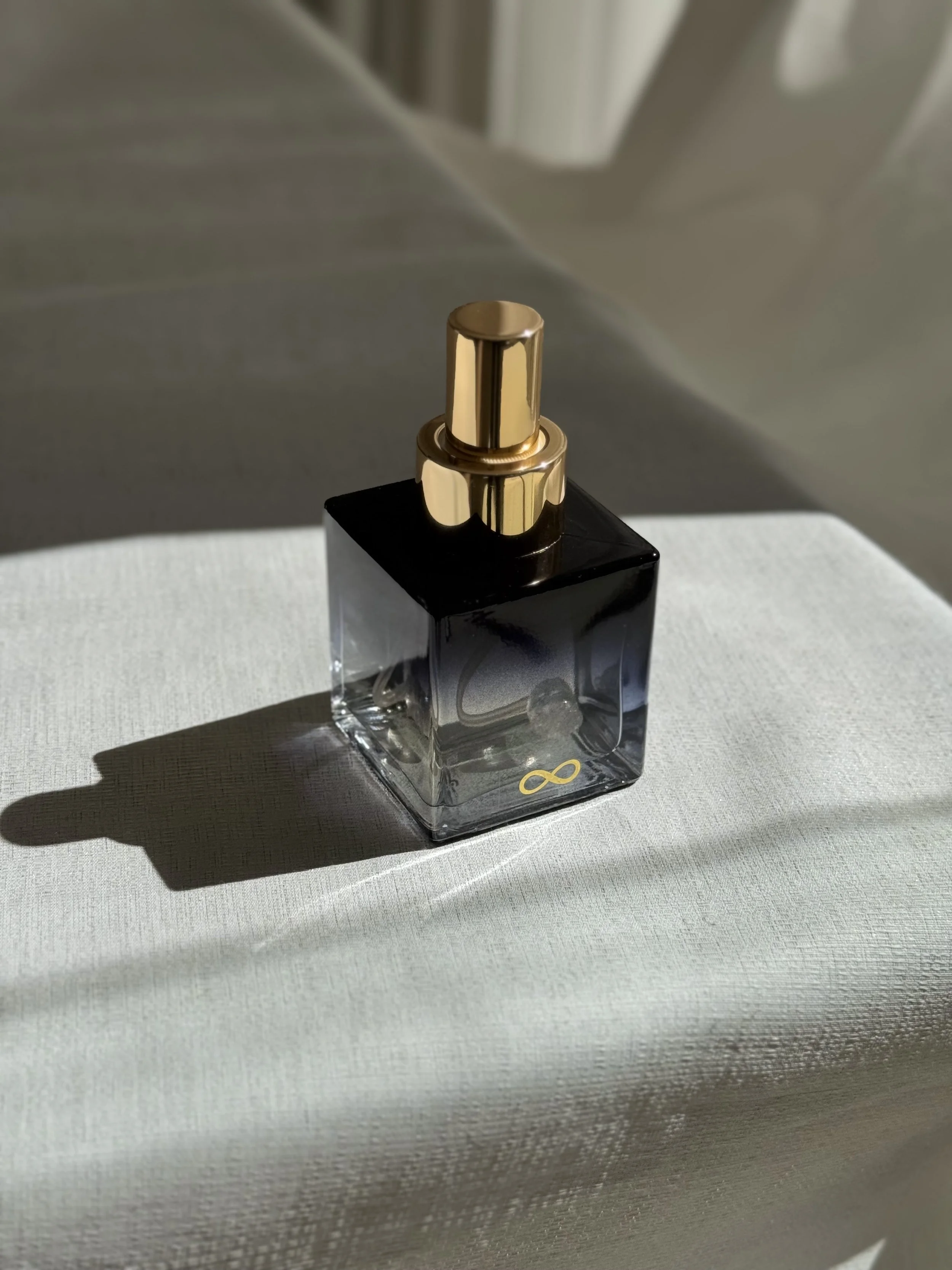 Close-up of a blue bespoke botanical perfume bottle with a gold cap, placed on a white textured surface with shadows cast to the left.