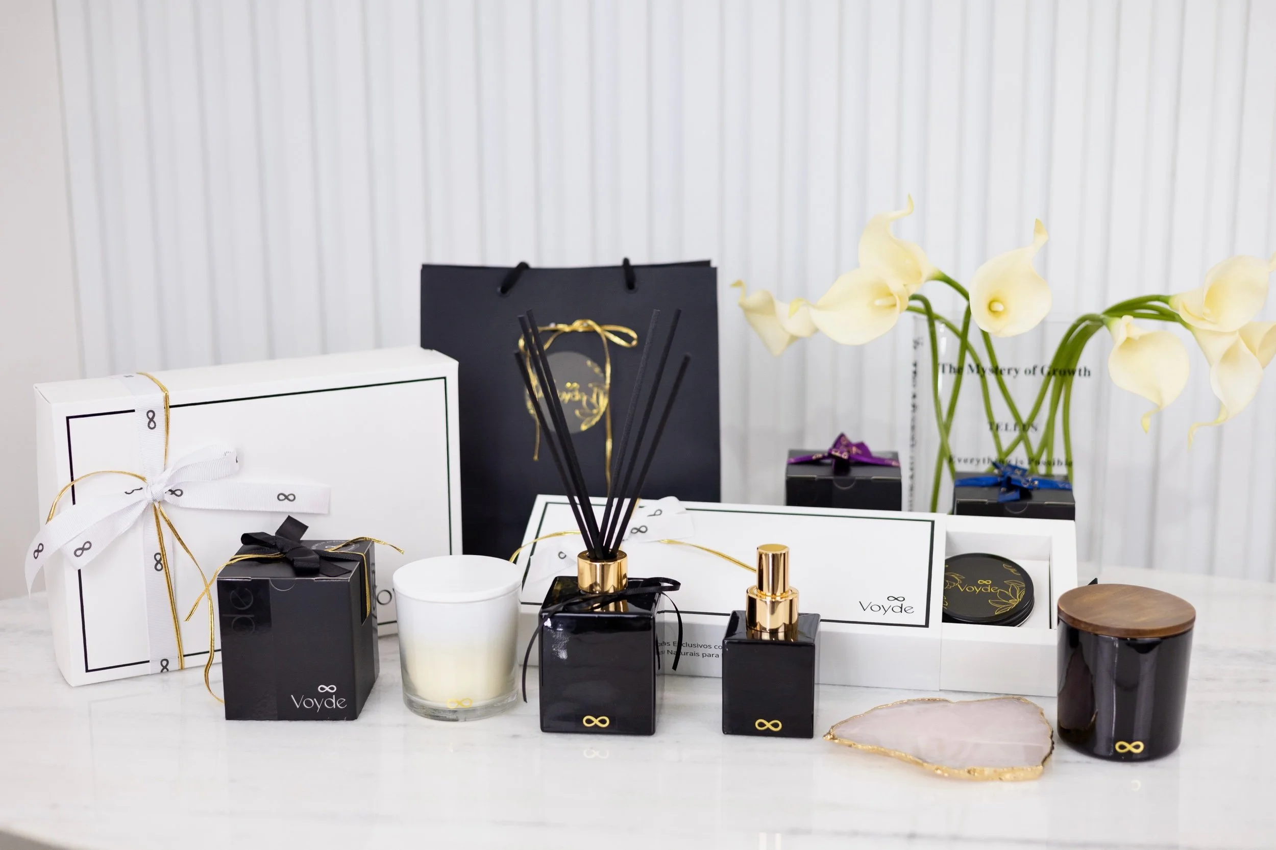 Display of skincare products and gifts, including a white box with a ribbon, black and white boxes, a white candle, perfume bottles, and a tray with flowers, against a white background with vertical lines.