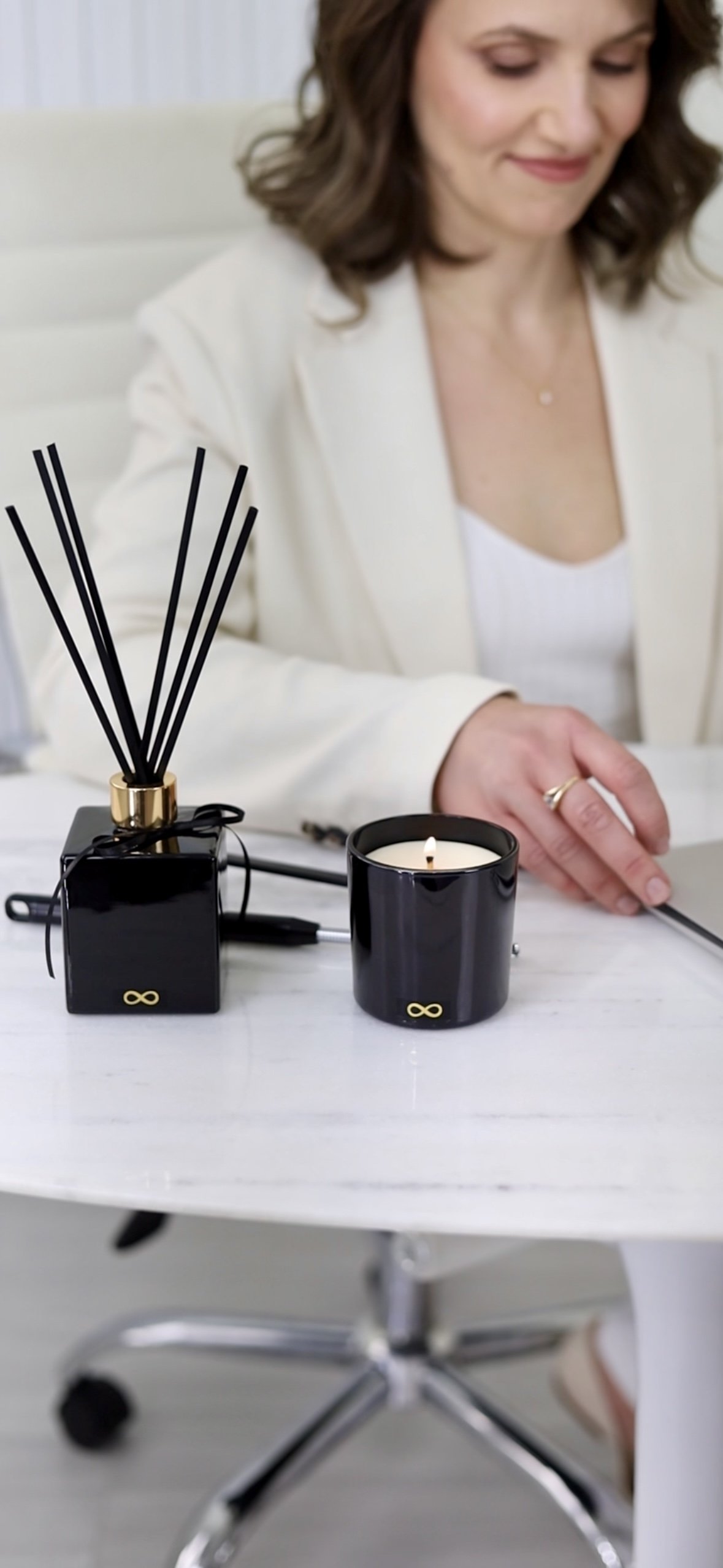 A elegant woman in a white jacket sitting at a white desk with a Voyde black botanical candle, a reed diffuser, and a silver pen.