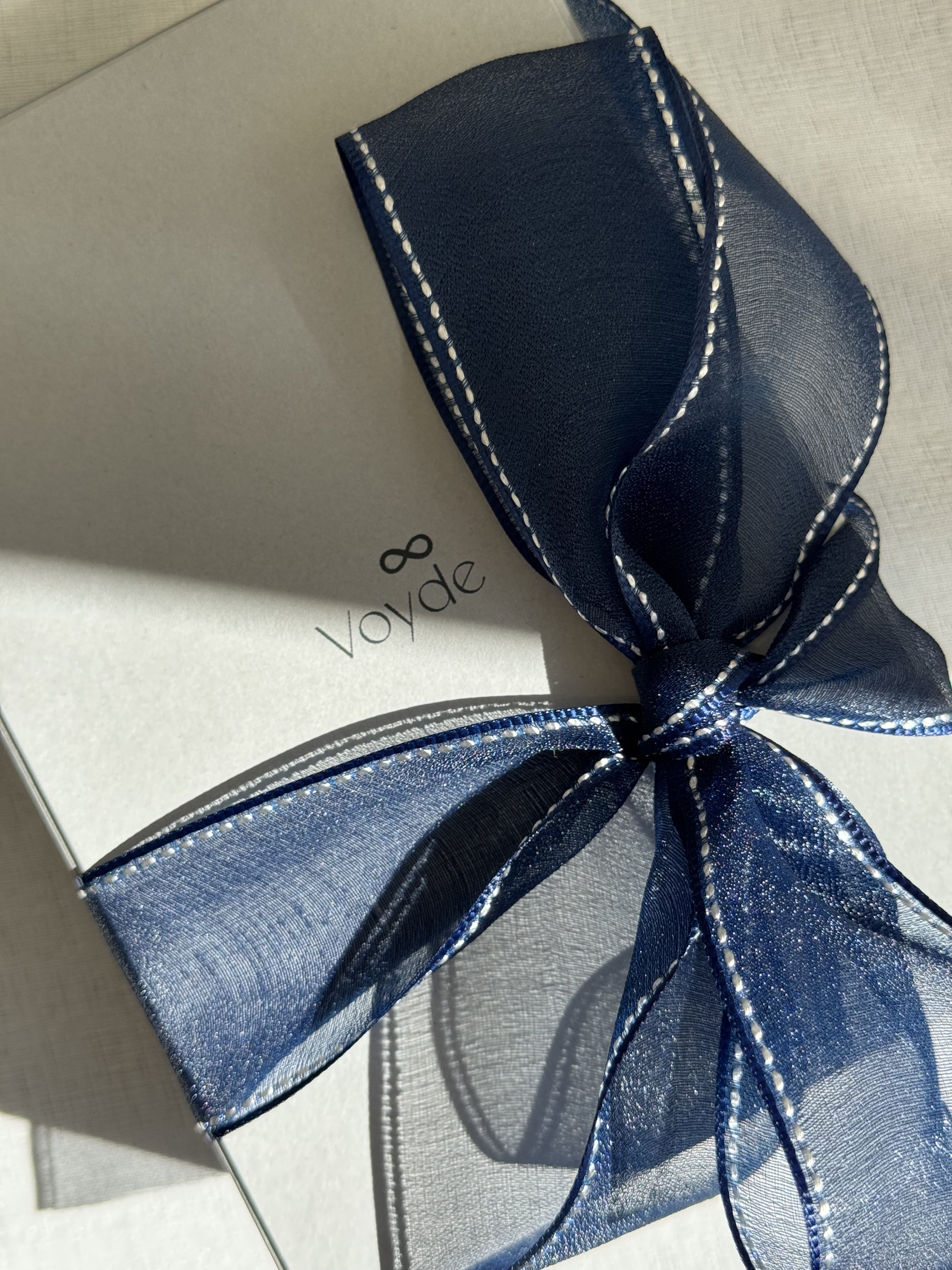 A light grey Voyde Bespoke Atelier box with a navy blue ribbon tied around it, resting on a reflective surface. The ribbon has white stitching along the edges and a metallic shimmer.