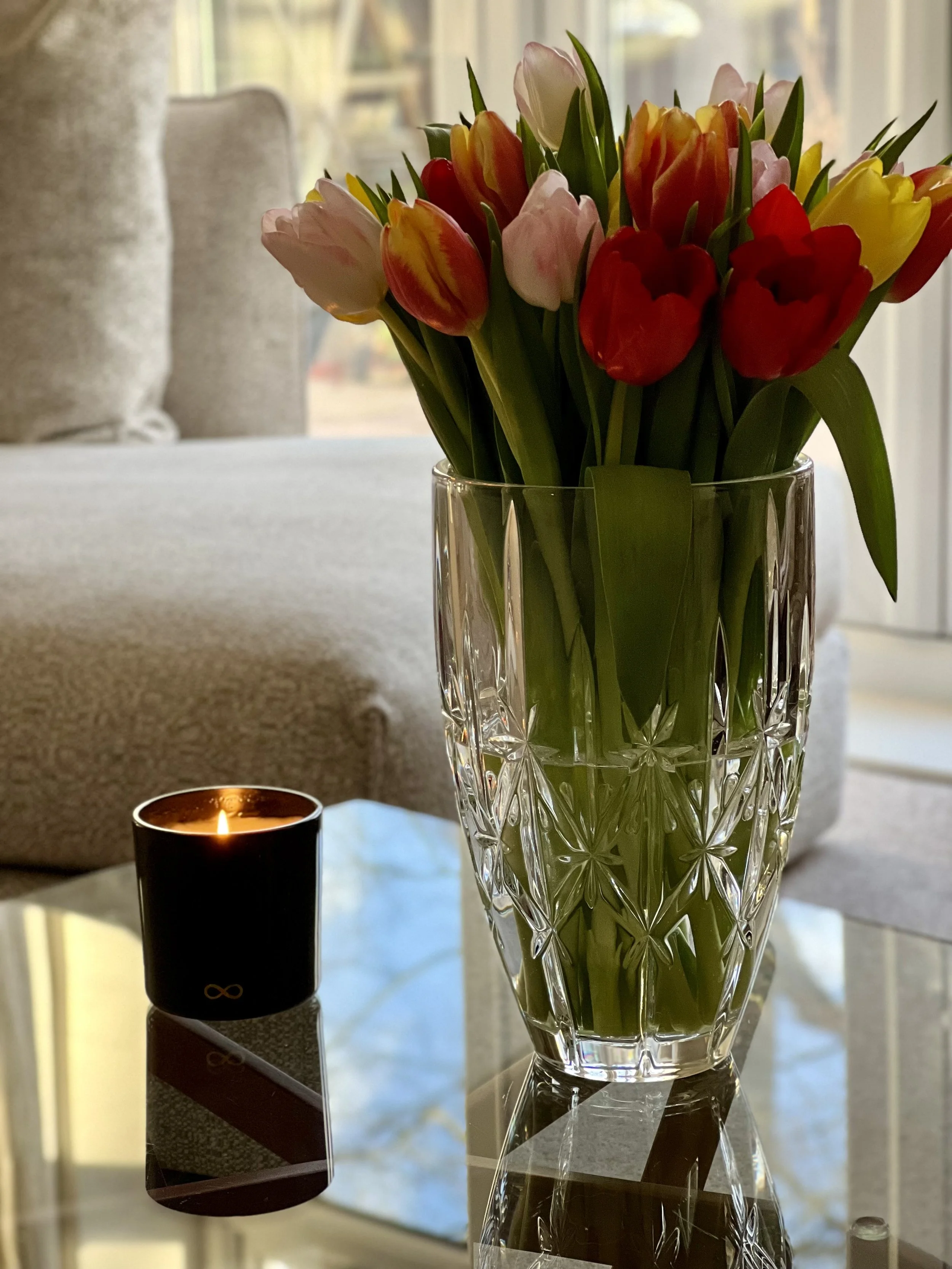 A clear glass vase with ornate patterns holds a bouquet of tulips in colors pink, red, and yellow, on a reflective surface with a lit black tealight candle nearby. In the background, there is a blurred view of a living room with light-colored furniture and large windows.