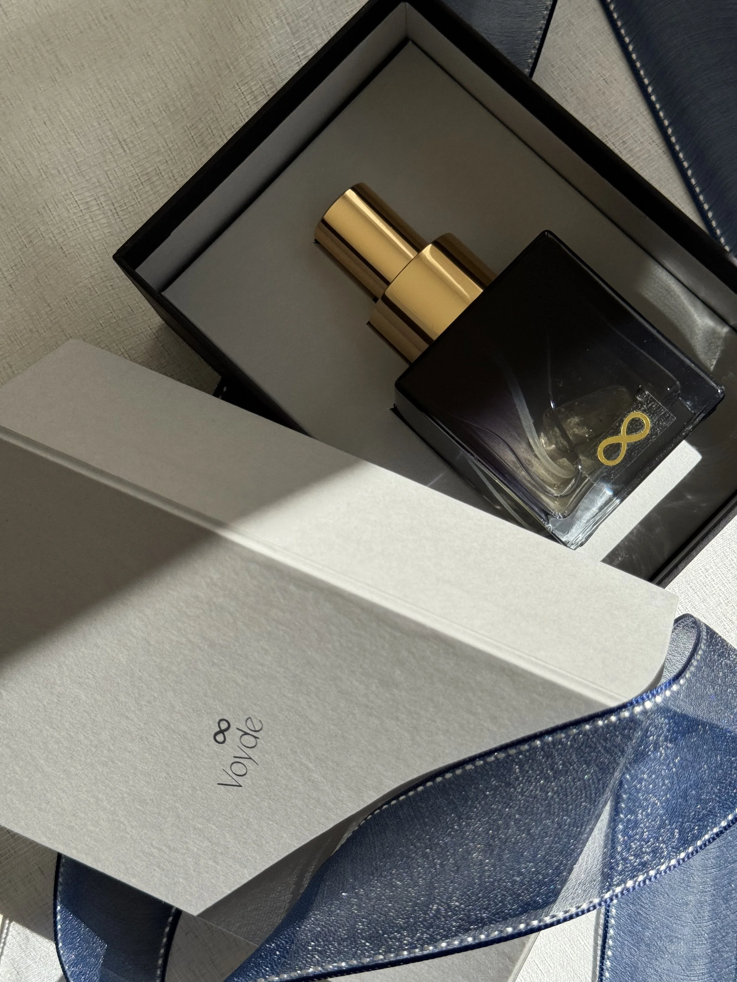 A gift box with a Voyde Bespoke perfum inside a blue elegant glass, and a blue textured ribbon beneath it.