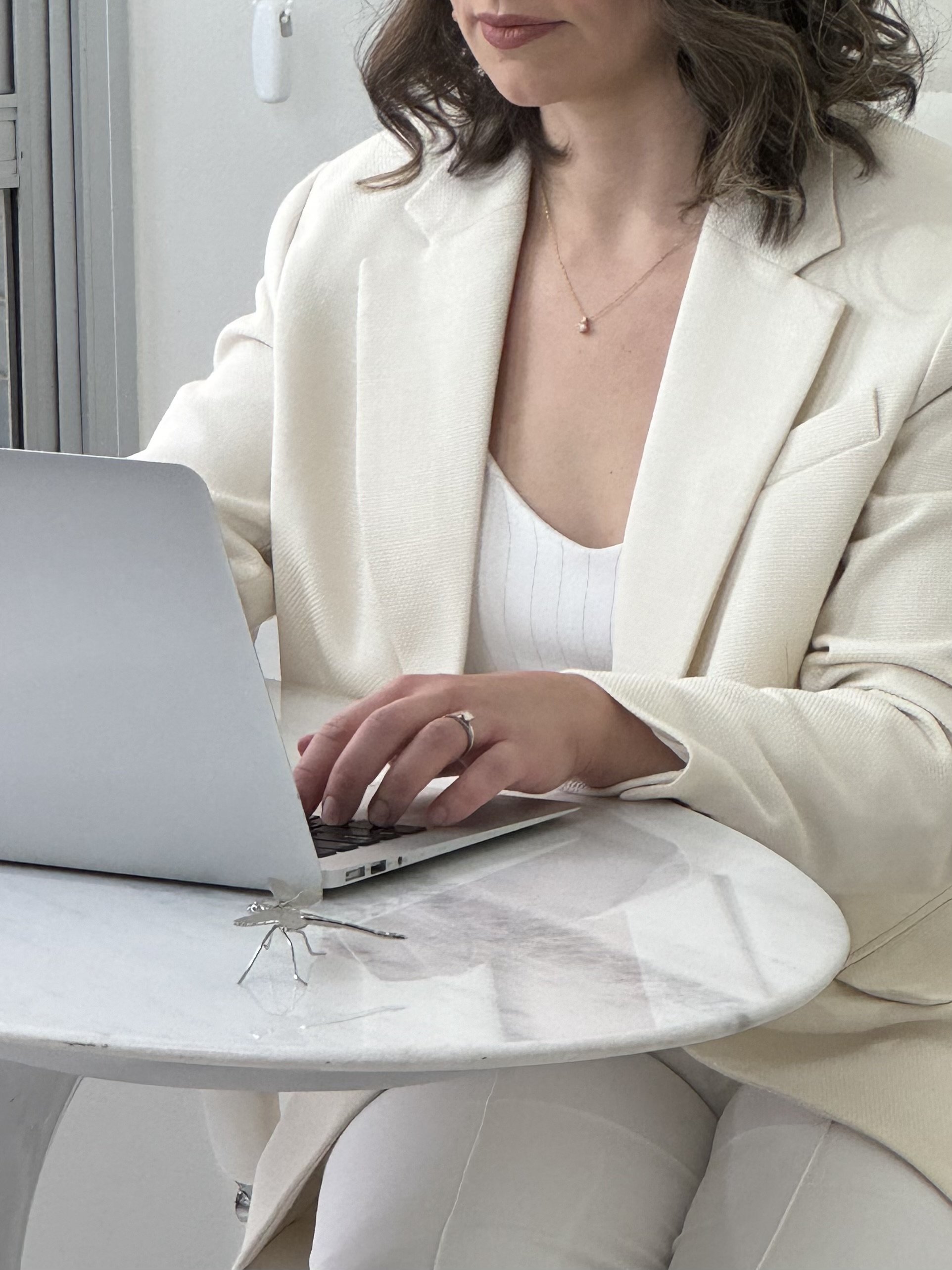 A elegant woman, wearing a white blazer and a matching top, is working on a laptop at a white table.