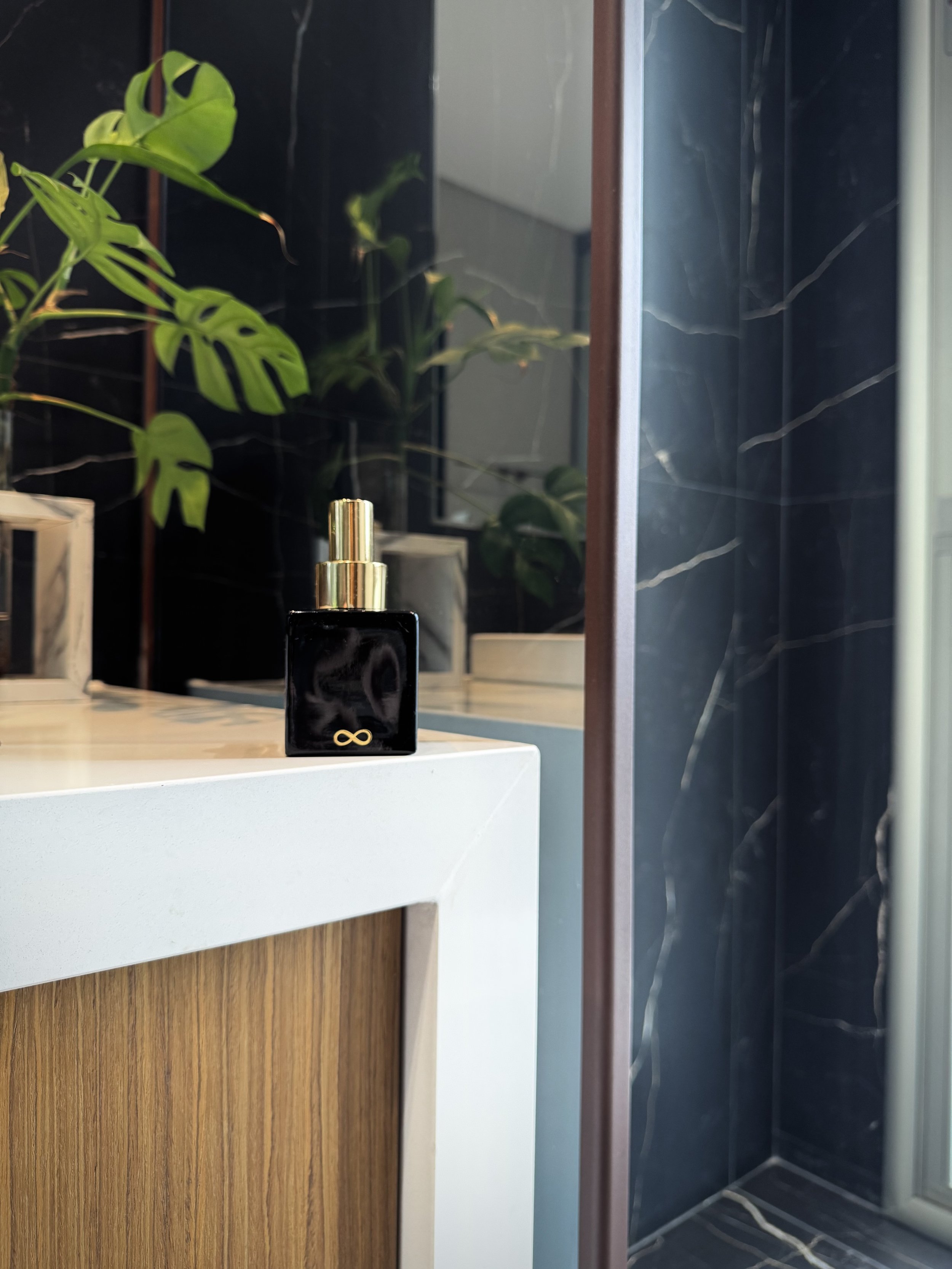 A bathroom vanity with a black energy mist spray bottle with a gold infinity symbol. There are green plants and mirror reflections visible in the background, along with black marble tiles with white veining.
