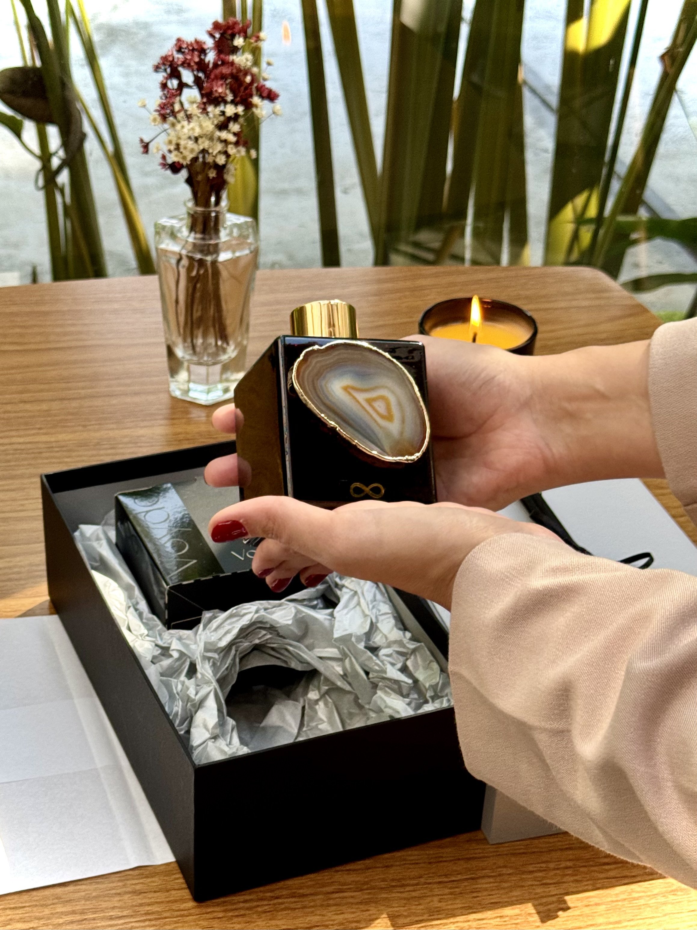 A person holding a reed diffuser with a large agate slice on top inside a black box with white tissue paper, a botaical candle on a wooden table.