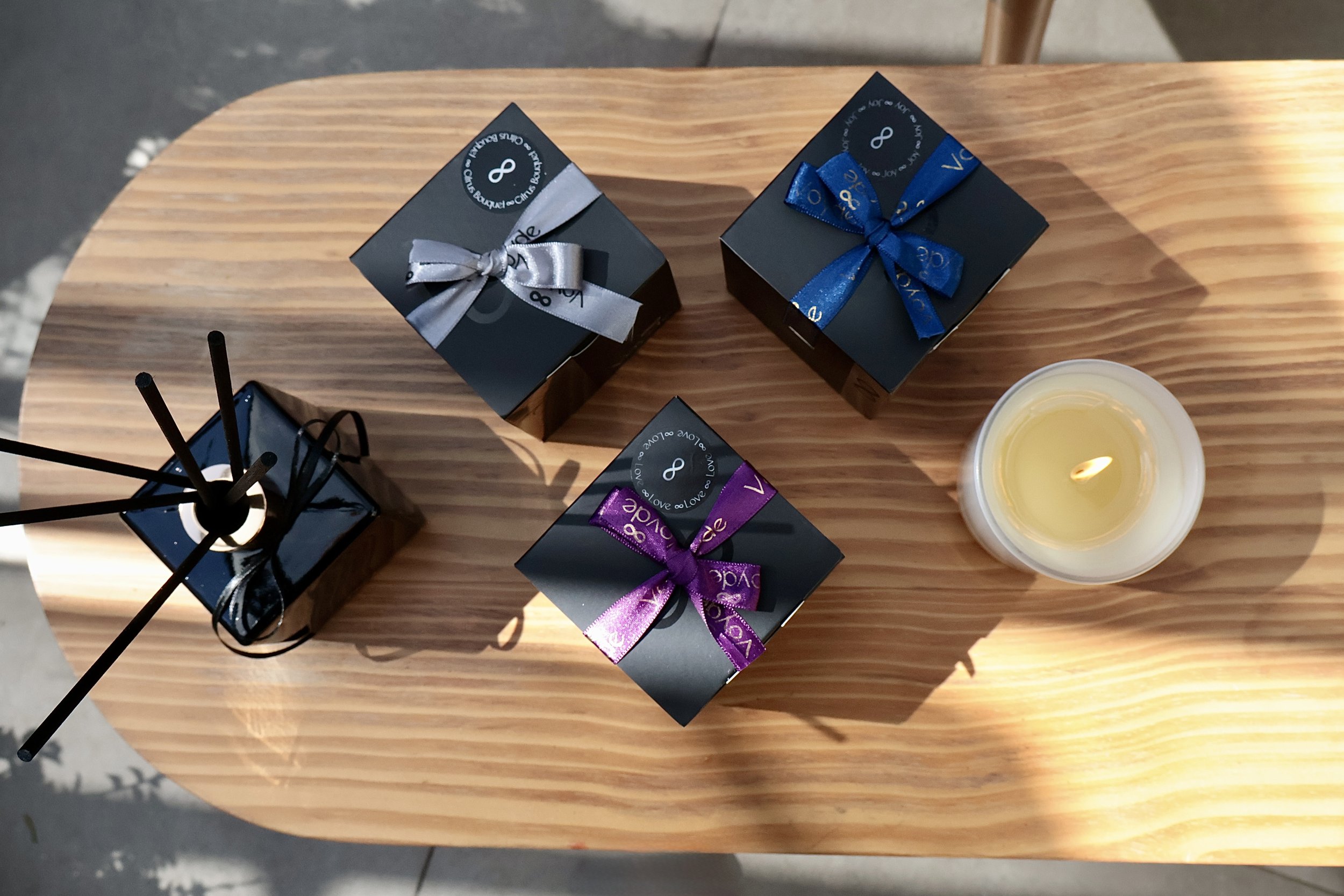 Three small black gift boxes with colorful ribbon bows on a light wooden table, alongside a lit white candle and a black reed diffuser.