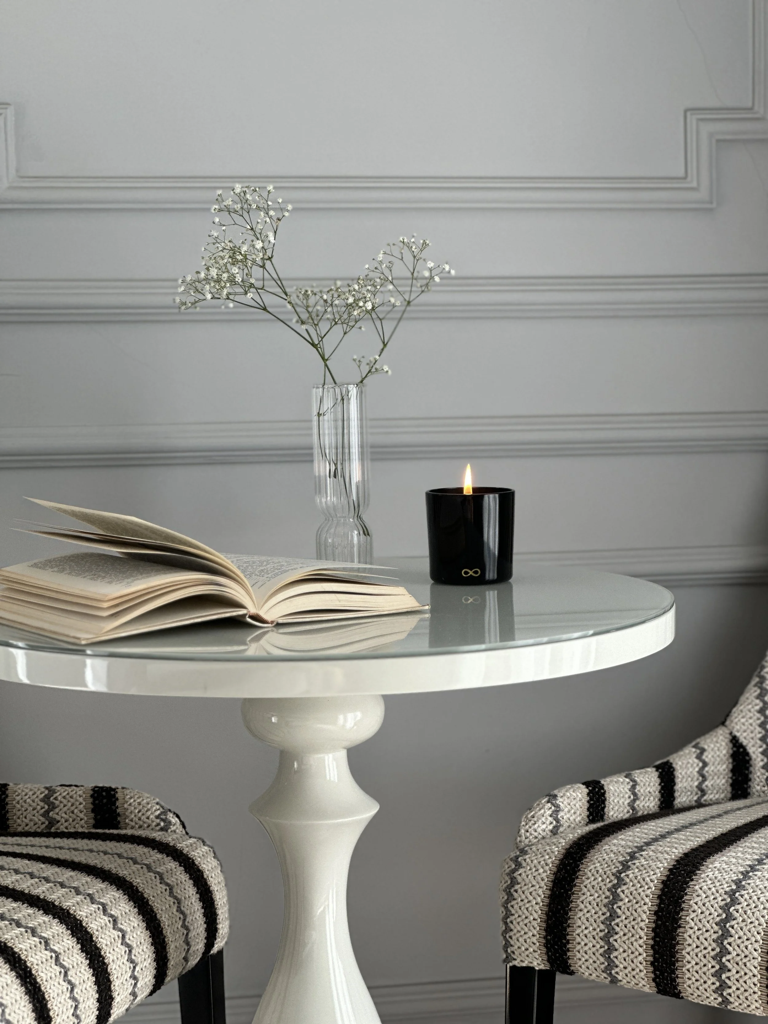 A white round table with a decorative pedestal base, a striped fabric chair on each side, a black candle holder with a lit candle, an open book, a glass vase with white baby's breath flowers, and a gray wall with decorative molding in the background.