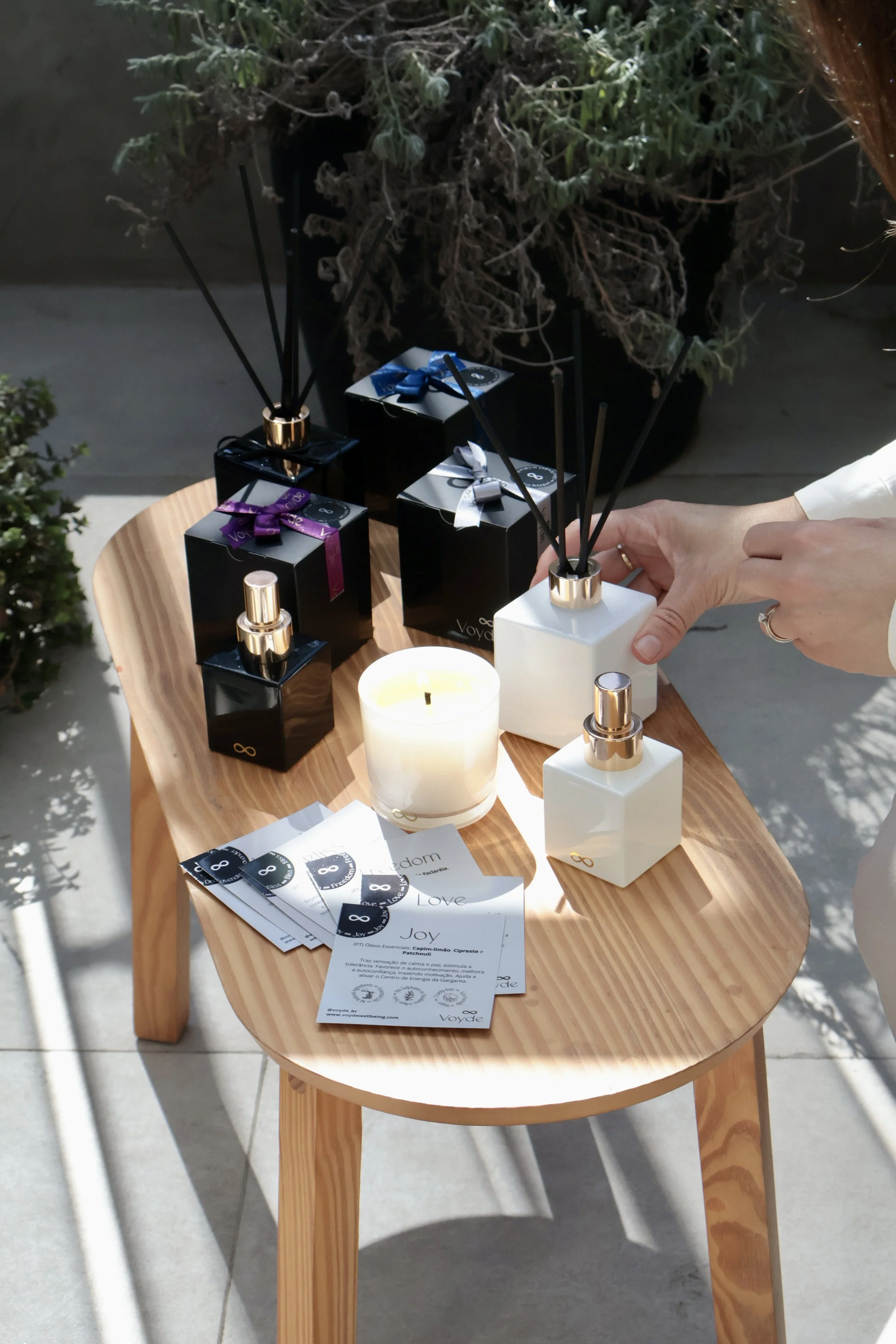 A wooden table displaying various black and white gift boxes with bows, botanical candles, energy mist, pillow mist spray and informational pamphlets, with a background of plants and sunlight.