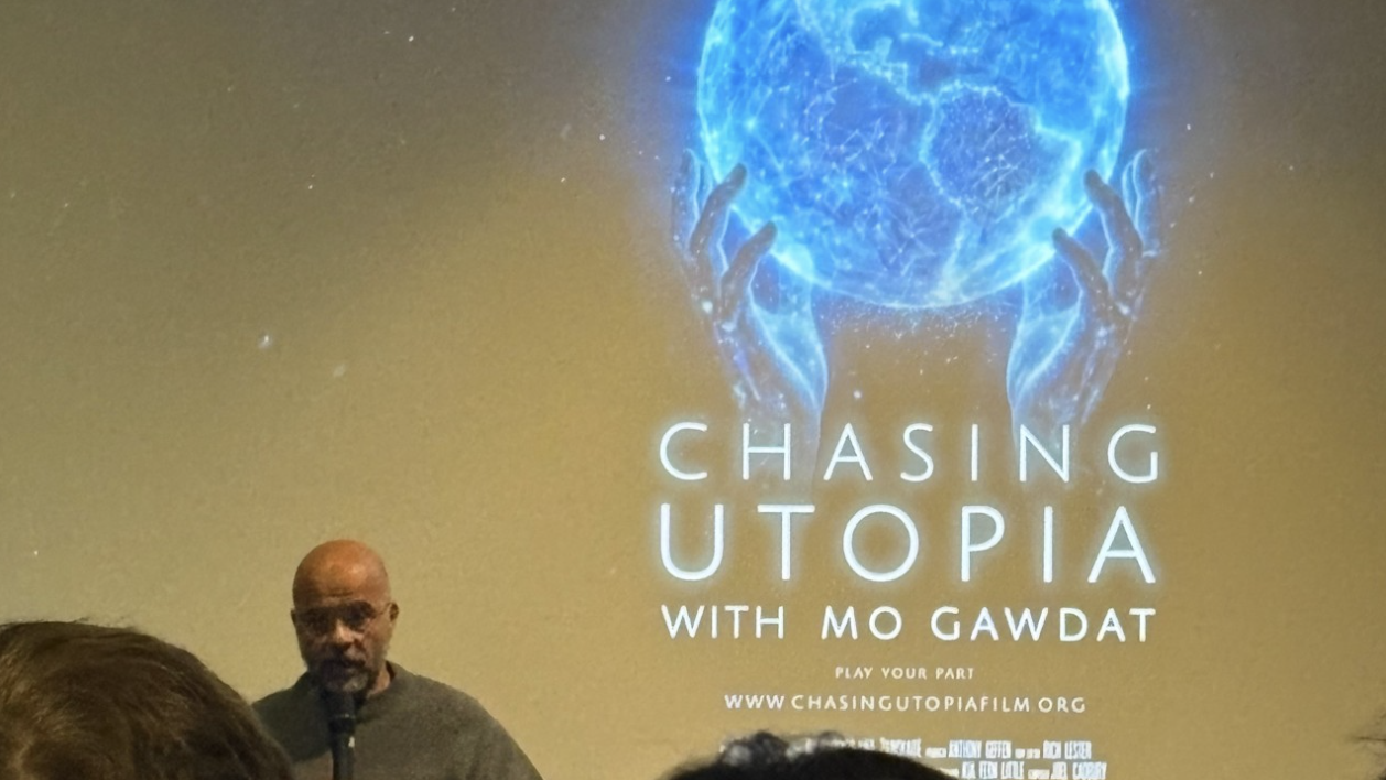 A man is speaking into a microphone in front of a projection that promotes the film 'Chasing Utopia' with Mo Gawdat, showing a pair of hands holding a glowing blue sphere.