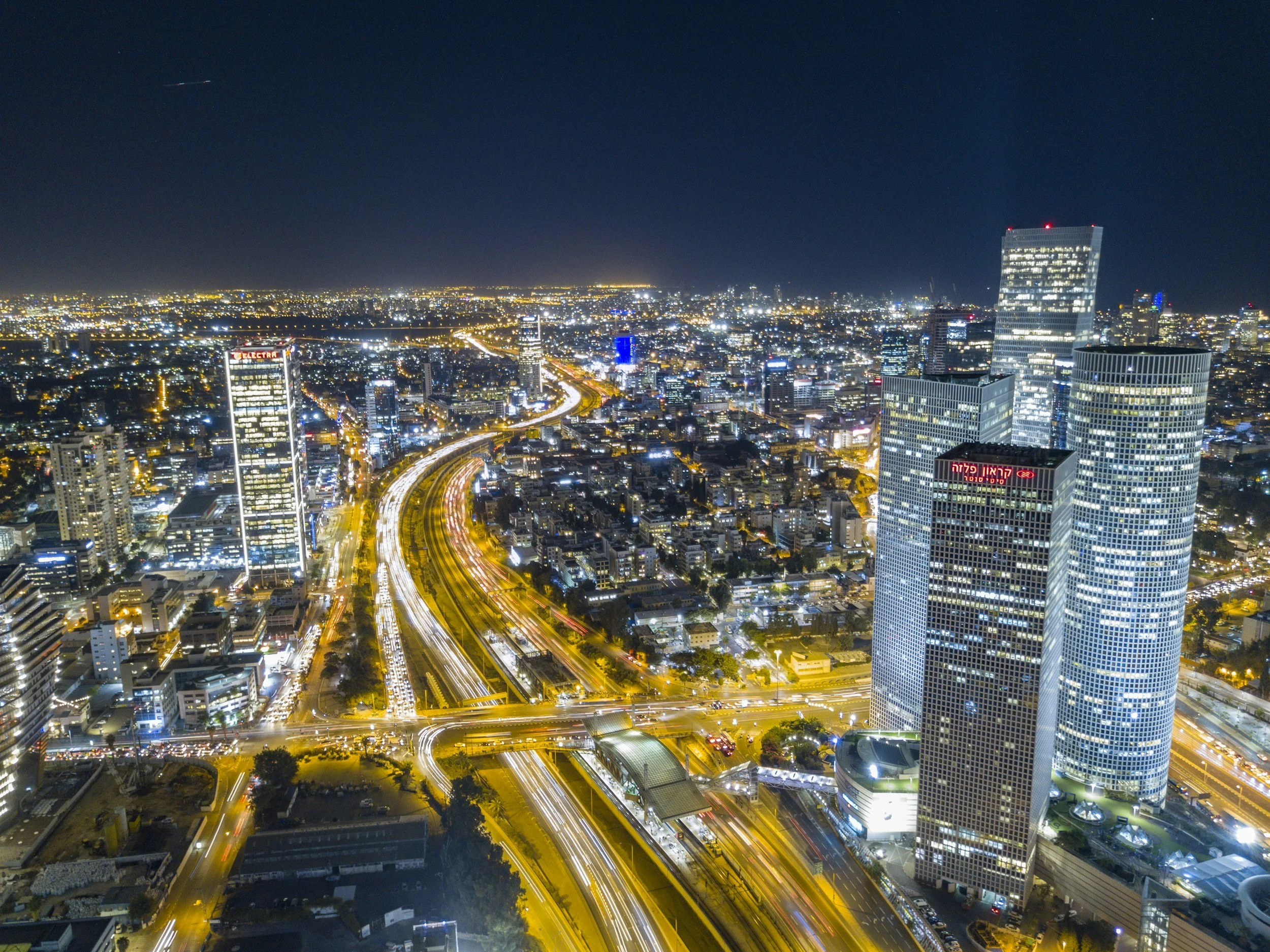 Nighttime aerial view of the buisness disrict of Tel Aviv and Ramat Gan in Israel including illuminated buildings, streets, and highways, including the Azrieli Centre, with a night sky overhead.