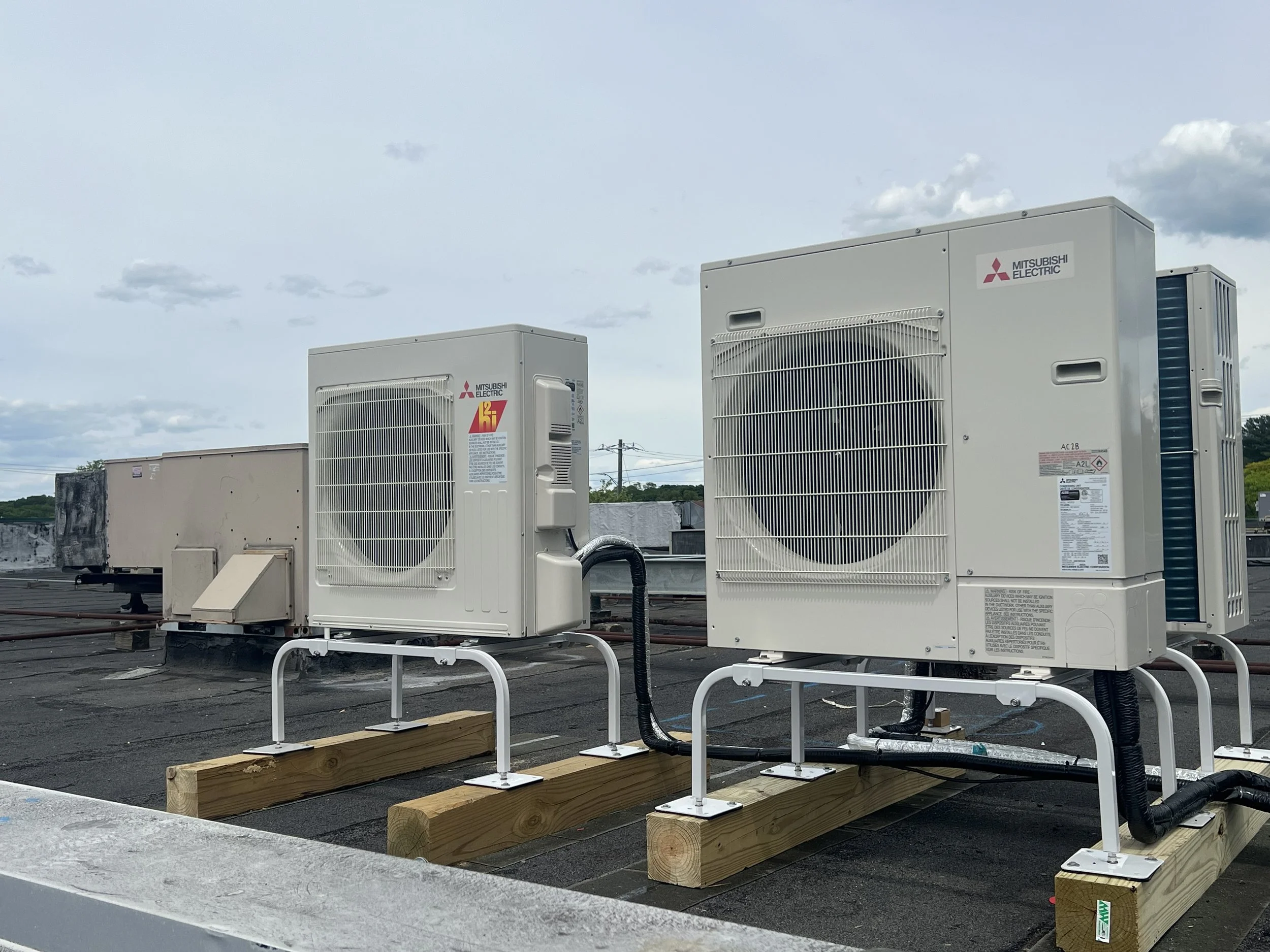 Several large Mitsubishi Electric HVAC units are installed on a rooftop with wooden blocks and metal rails for support, under a partly cloudy sky.