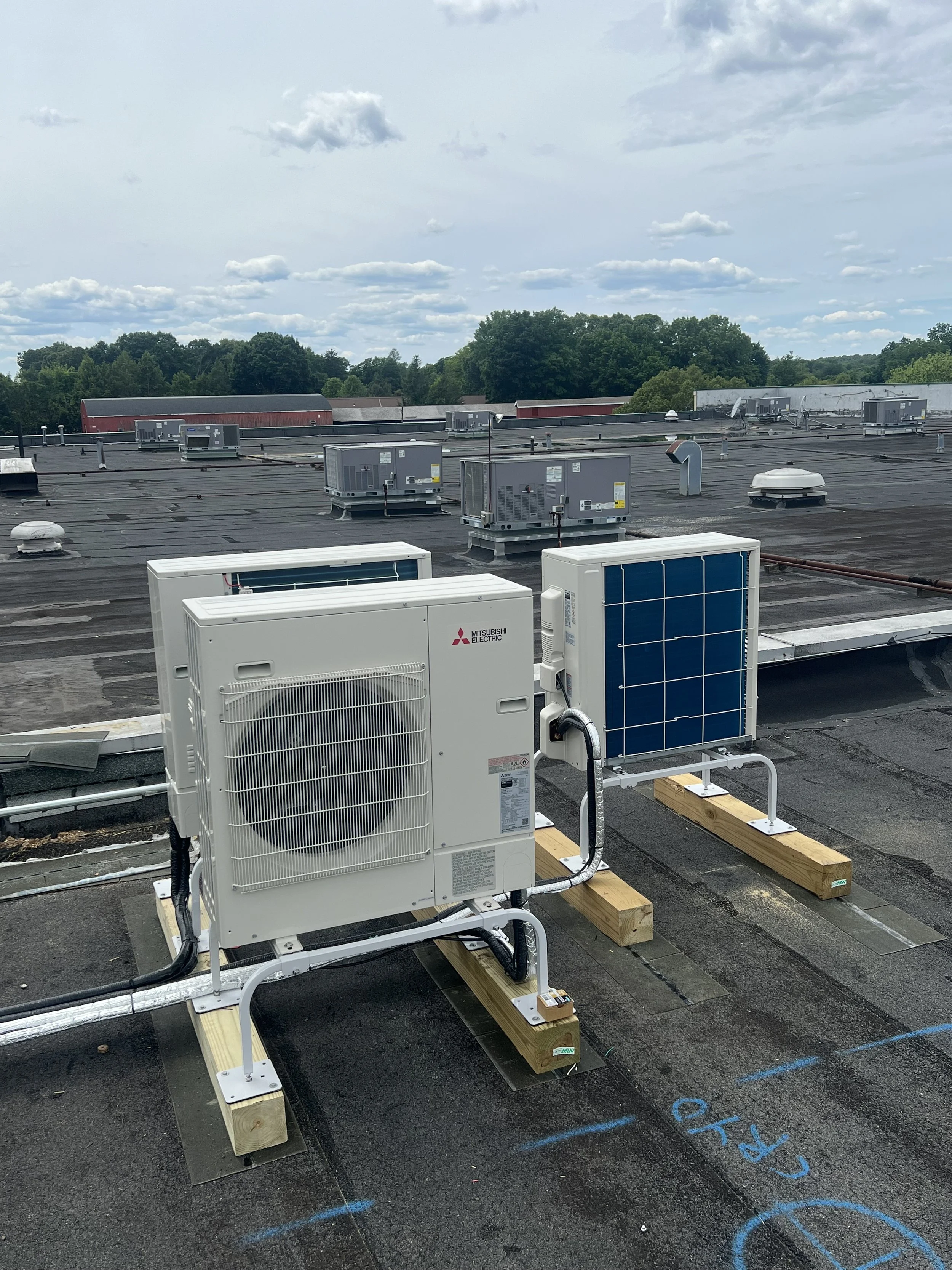 Rooftop with HVAC units and equipment, wooden support beams, and a view of green trees and a partly cloudy sky in the background.