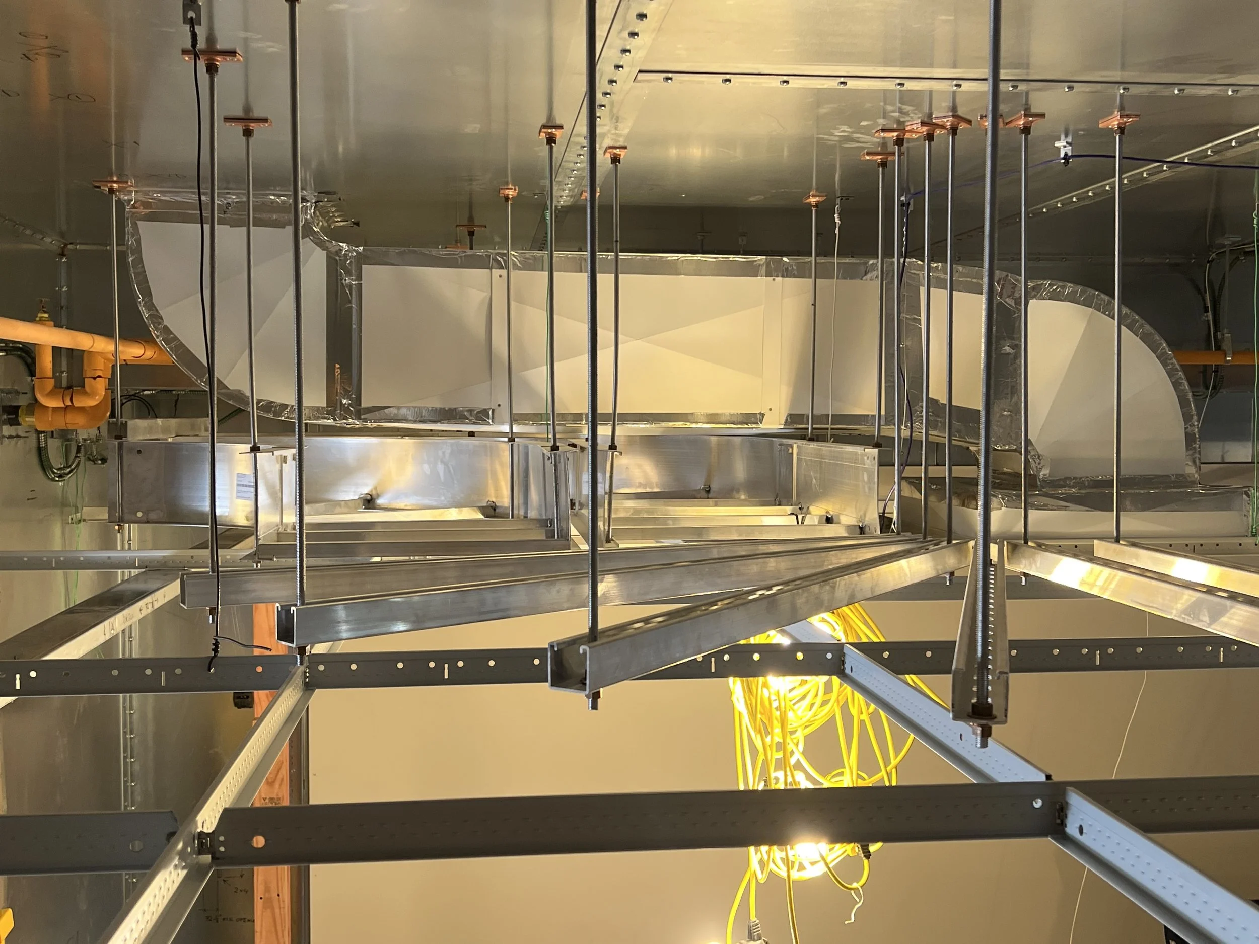 This image shows an unfinished ceiling in a building under construction, with metal framing, orange pipes, yellow electrical wiring, and some wooden supports.