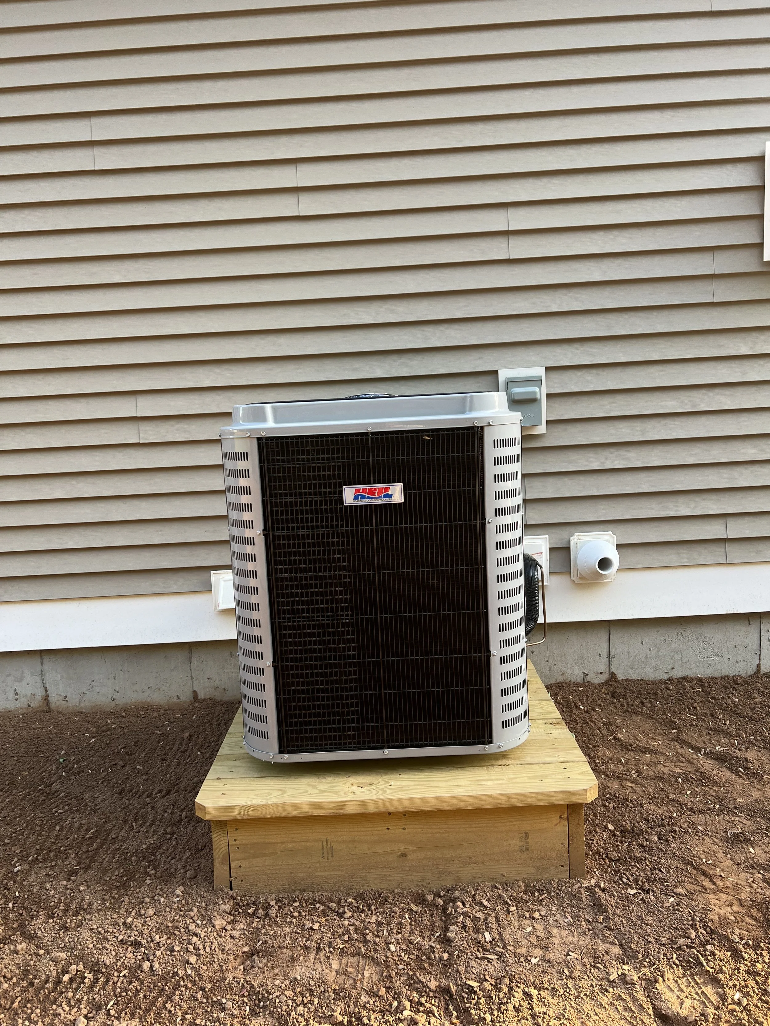 The image shows an air conditioning unit installed outdoors on a wooden platform, in front of a beige house siding wall. The ground around the platform is dirt, and there are electrical outlets and conduits on the wall.