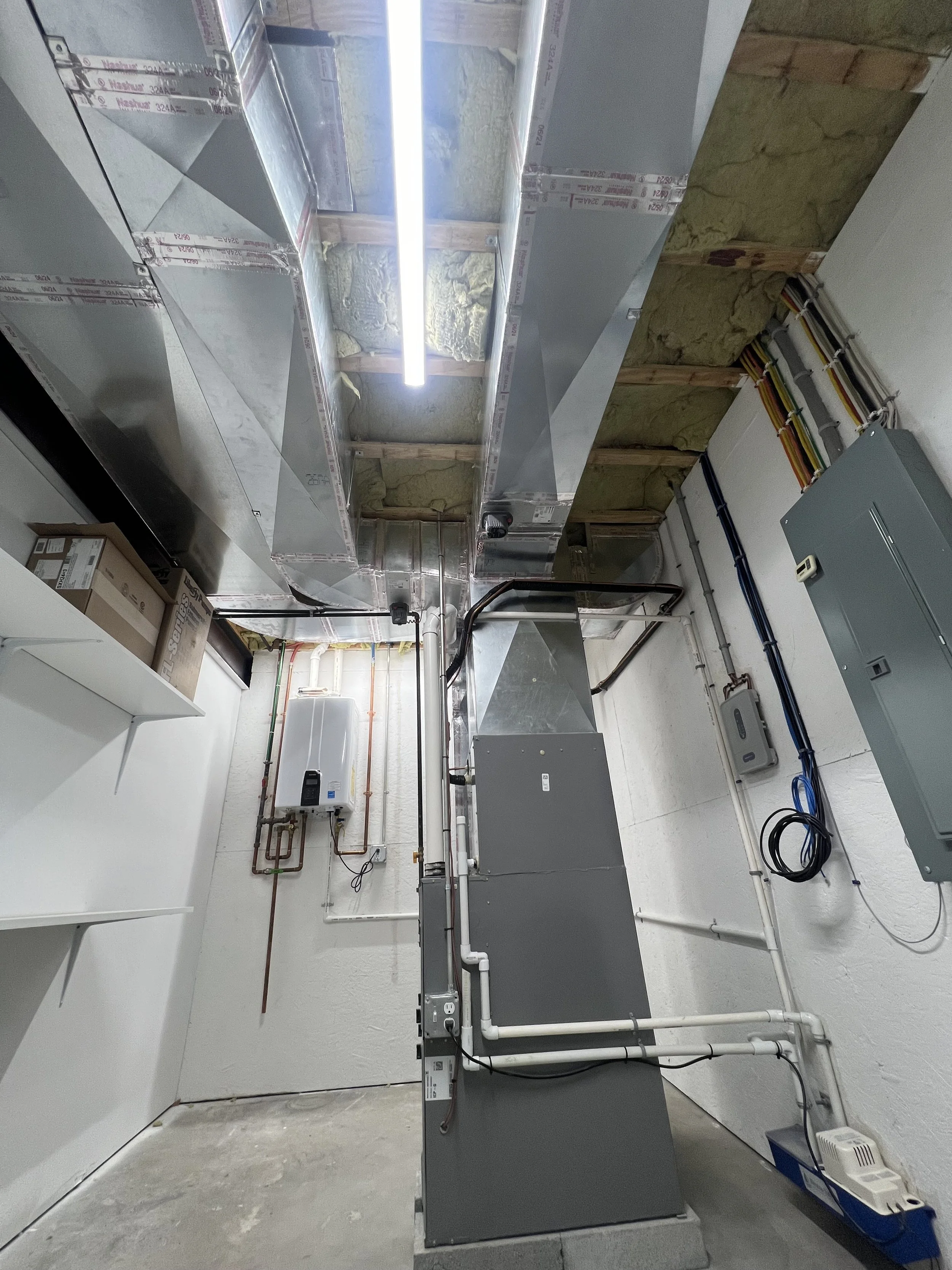 A utility or mechanical room with ductwork, electrical conduits, and pipes installed along the walls and ceiling, with insulation visible behind the ductwork and a fluorescent light fixture overhead.