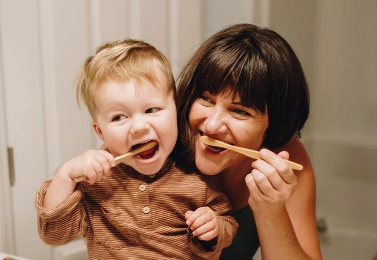 A woman and young boy with wood utensils in their mouths, pretending to eat. The woman is smiling and the boy is playful, indoors.
