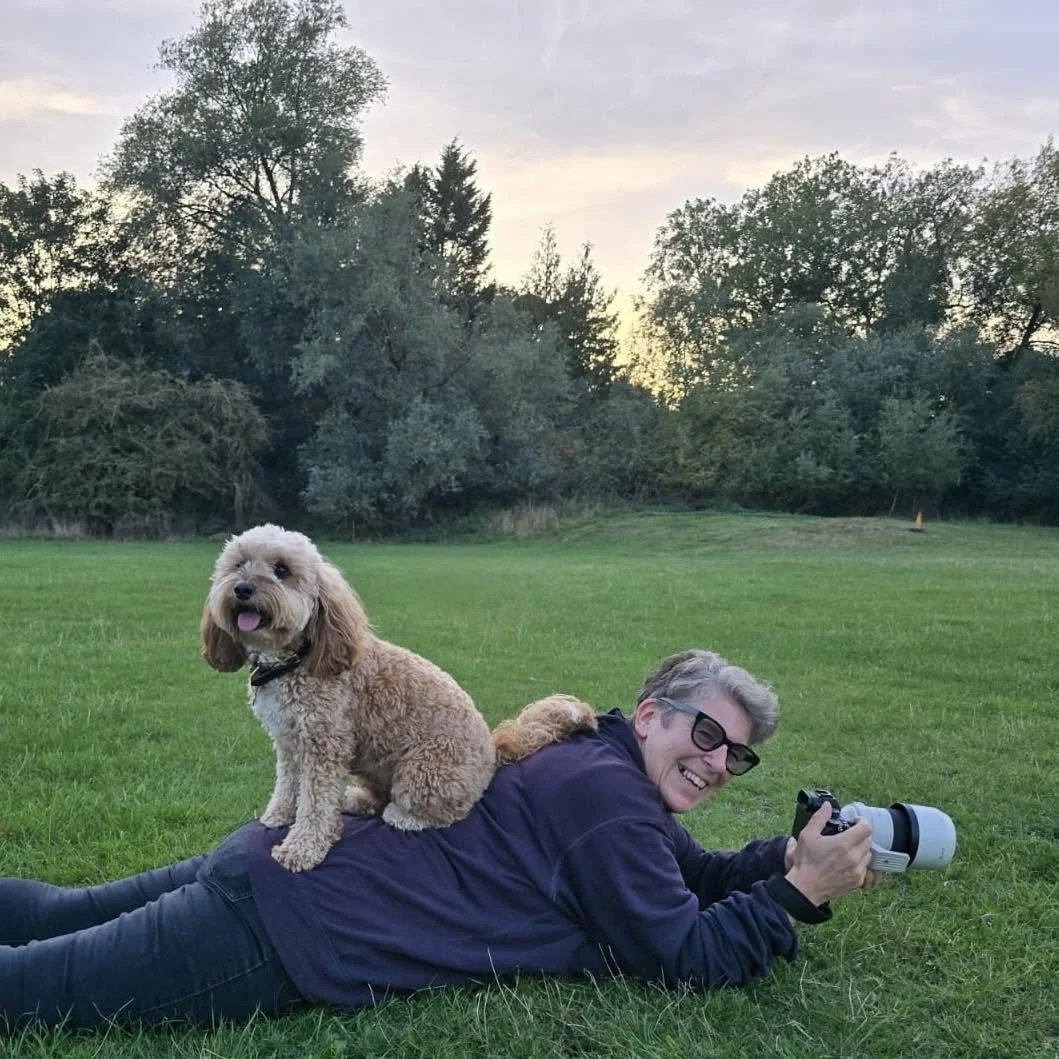 A man with glasses lying on the grass outdoors, smiling and holding a camera, with a light-colored curly-haired dog sitting on his back, in a park with trees and a cloudy sky in the background.