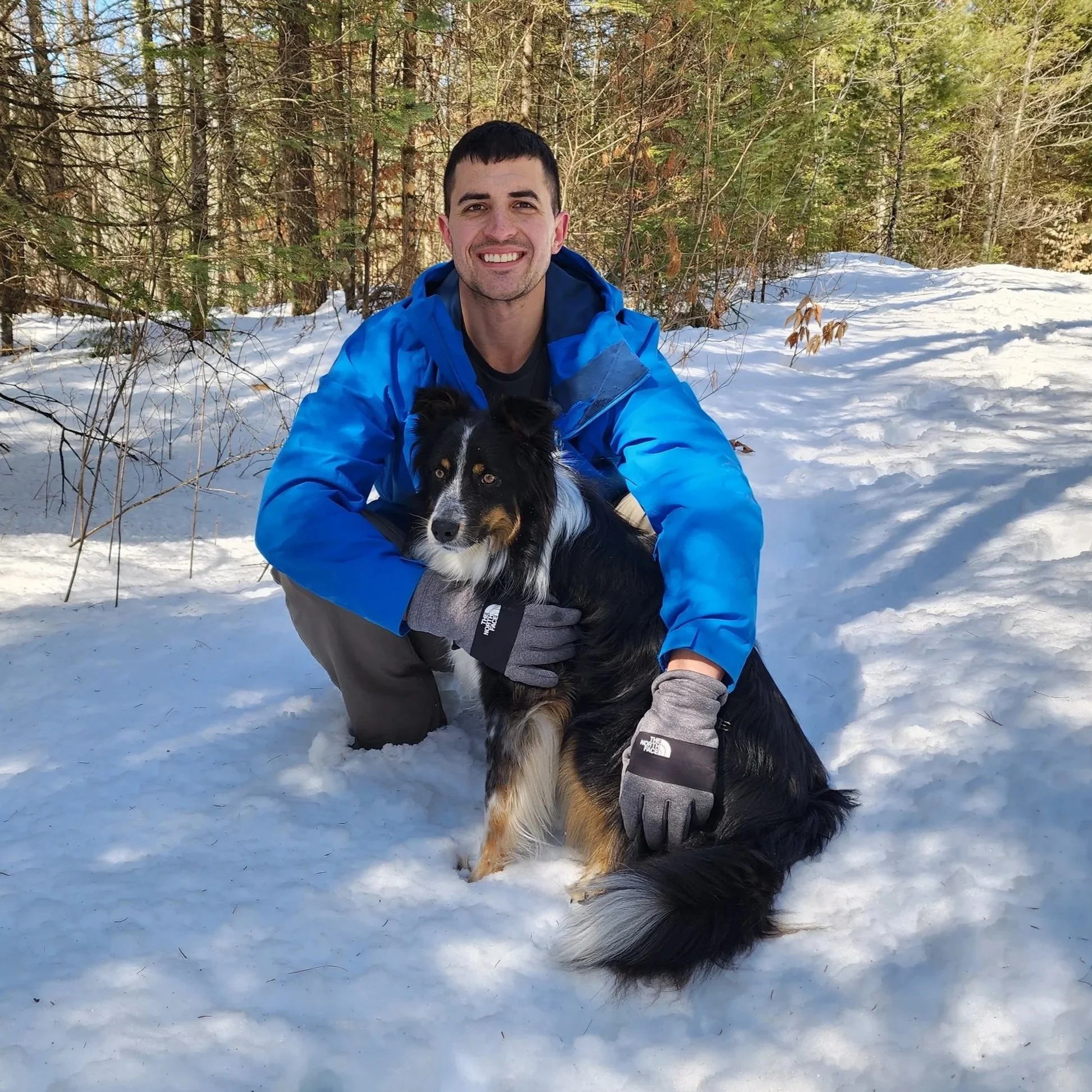 A man with short dark hair, wearing a blue jacket and gray gloves, kneeling in the snow outdoors, smiling at the camera, holding a black and white Australian Shepherd dog in a snowy forest.