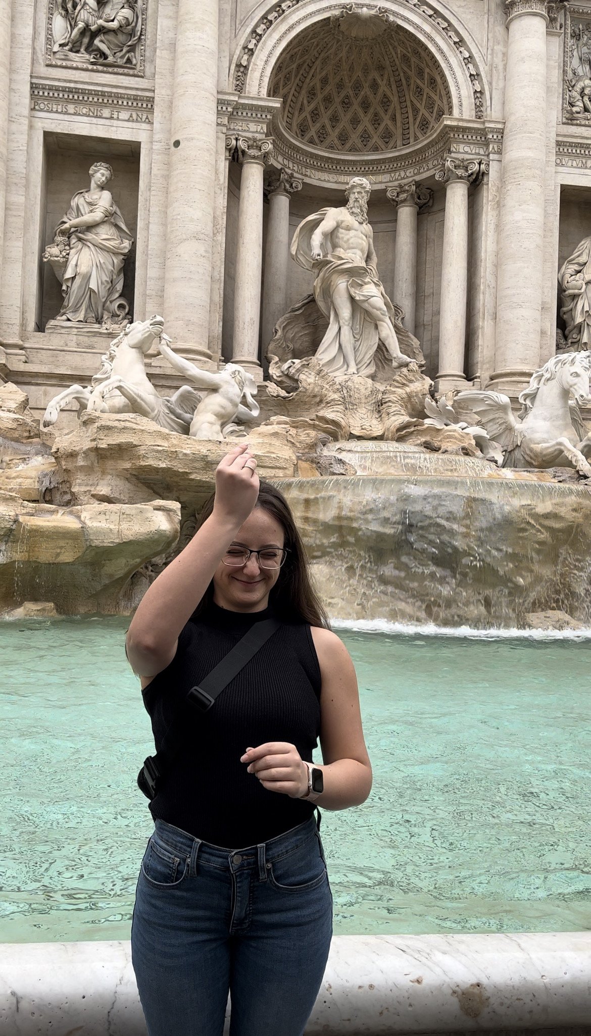 A woman standing in front of the Trevi Fountain in Rome, Italy, smiling and raising her right hand, with the ornate stone sculptures and water fountain as the backdrop.
