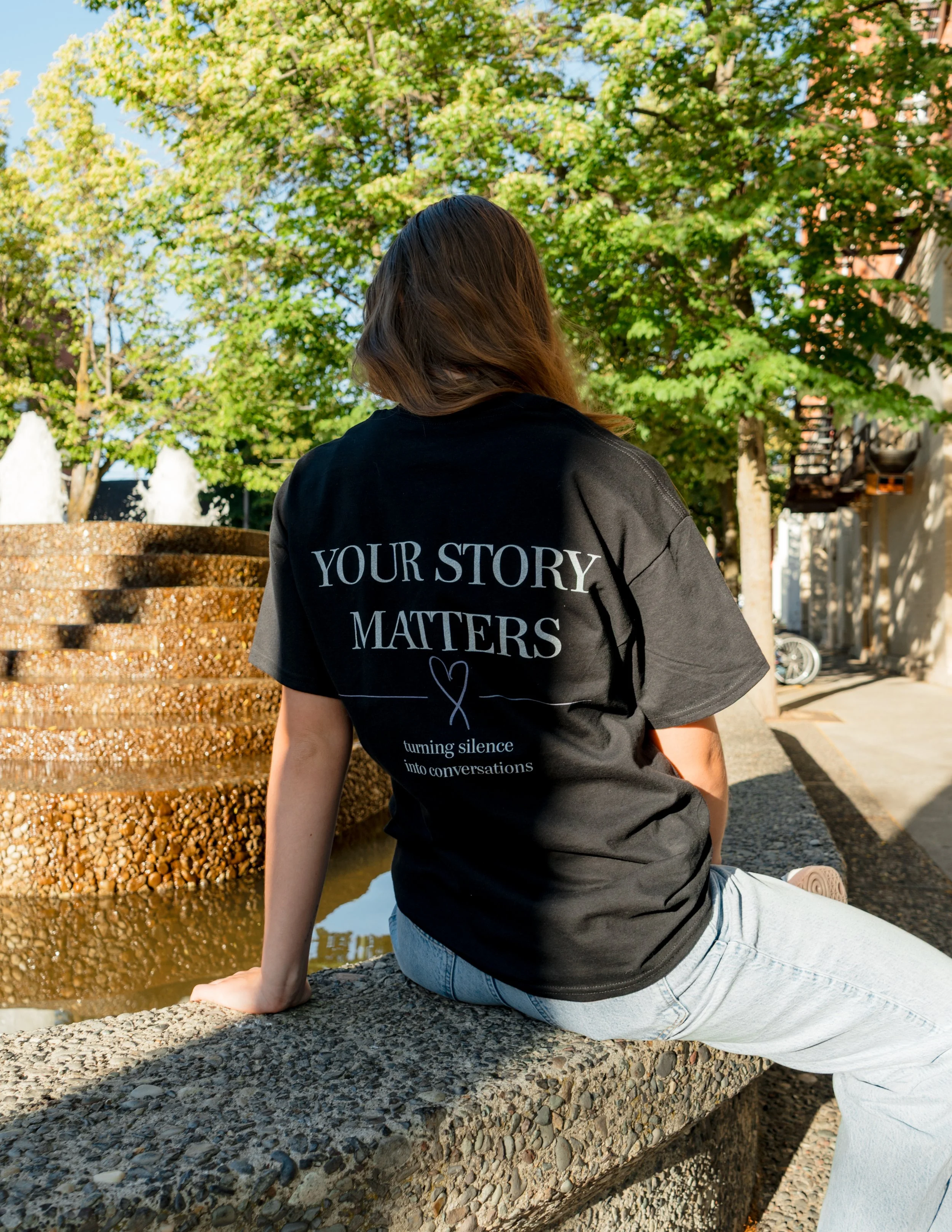 Woman wearing a StandUp33 "Your Story Matters" T-shirt while sitting near a fountain.