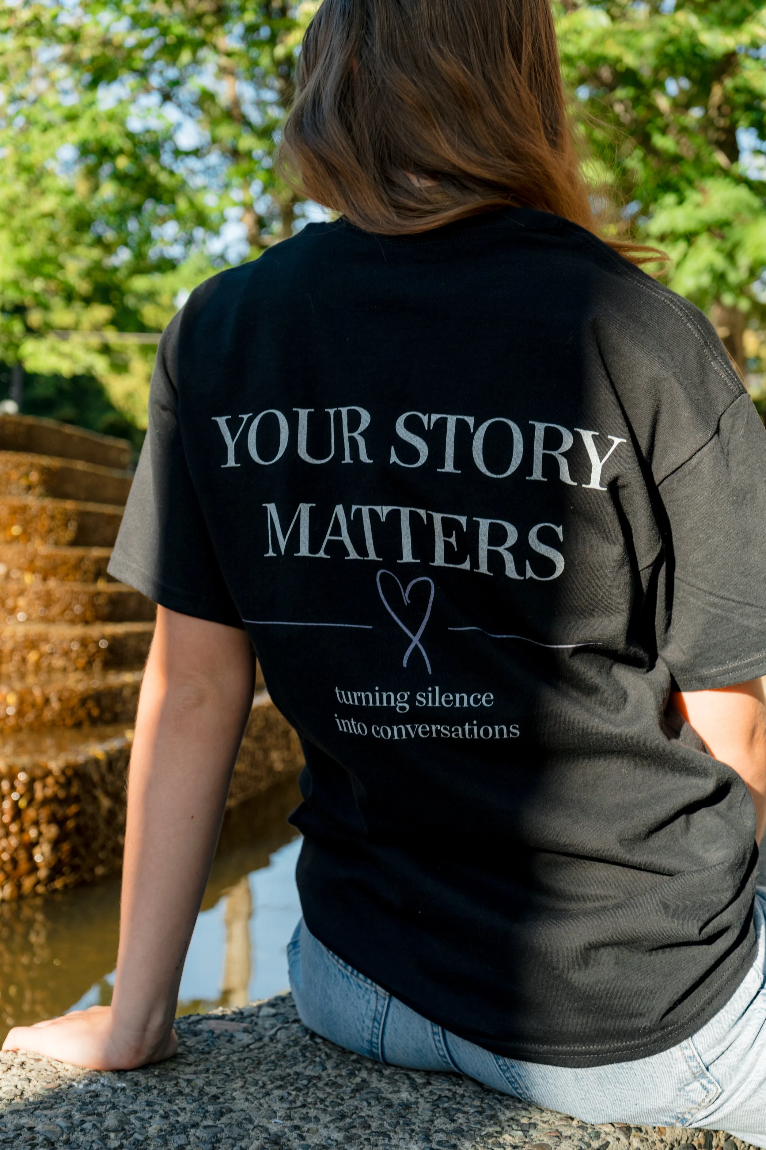 Back view of a woman wearing a StandUp33 "Your Story Matters" T-shirt while sitting near a fountain.