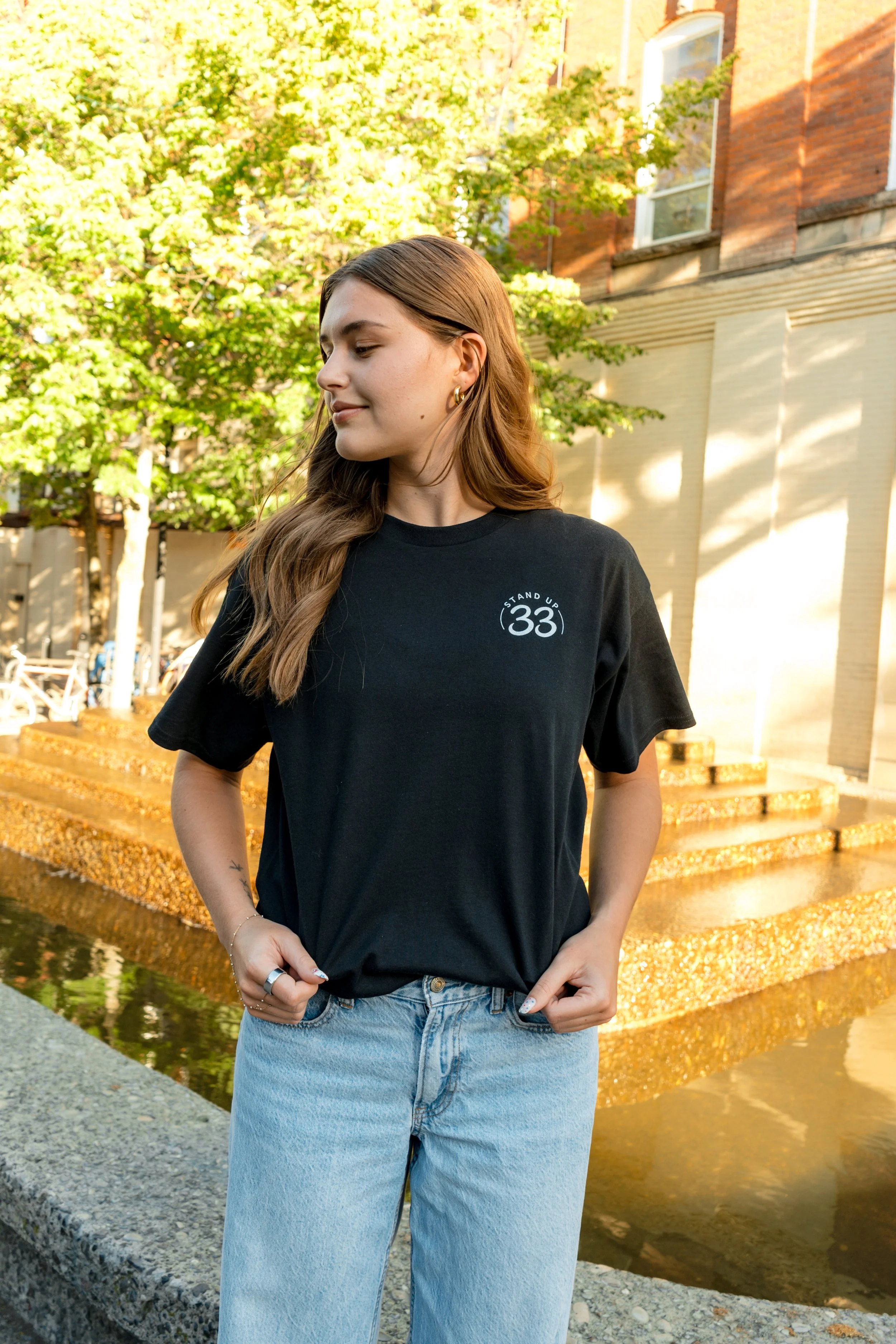 Woman wearing a StandUp33 logo T-shirt while standing outdoors near a fountain.