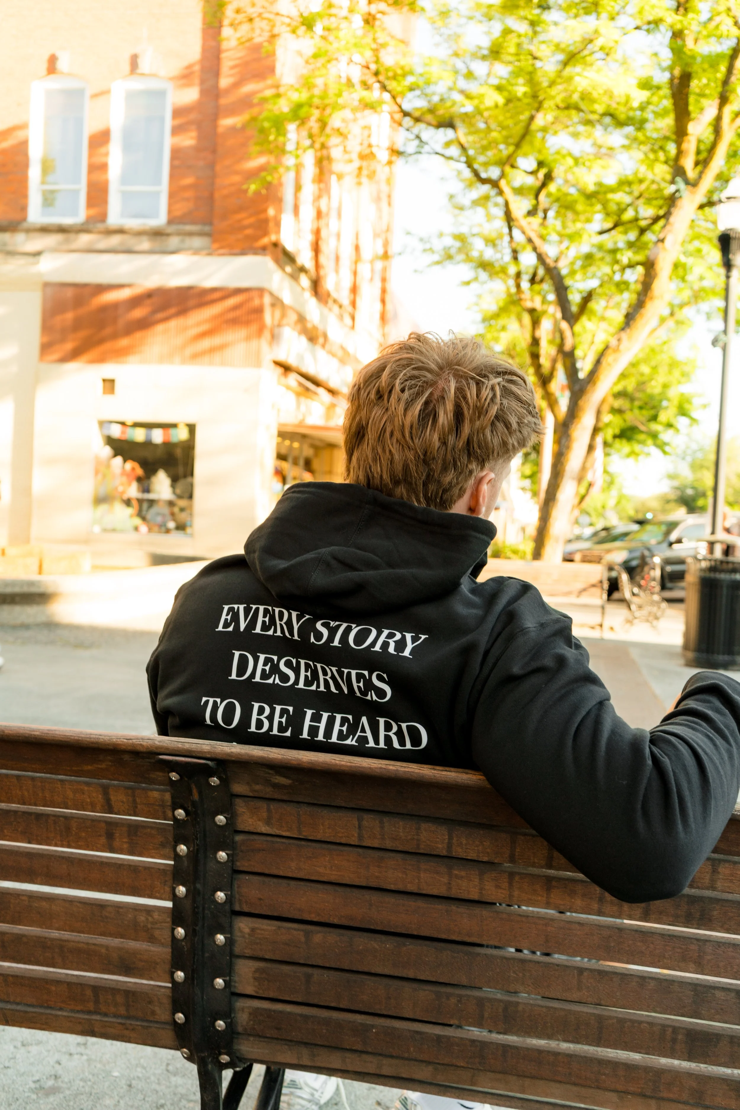 Back view of a person wearing a StandUp33 hoodie with the message "Every Story Deserves to Be Heard" while sitting on a park bench.