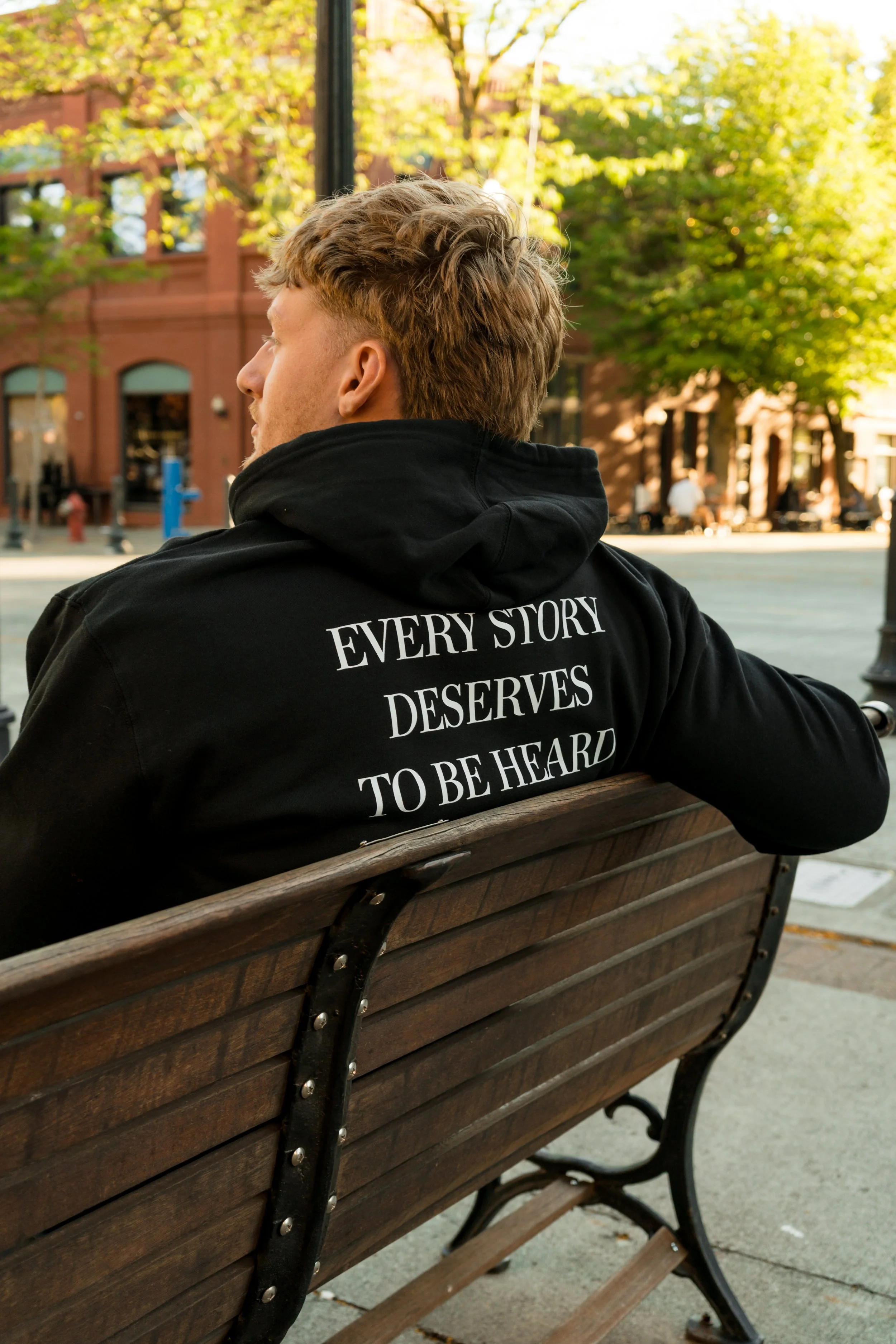 Person wearing a StandUp33 hoodie with the message "Every Story Deserves to Be Heard" while sitting on a park bench.