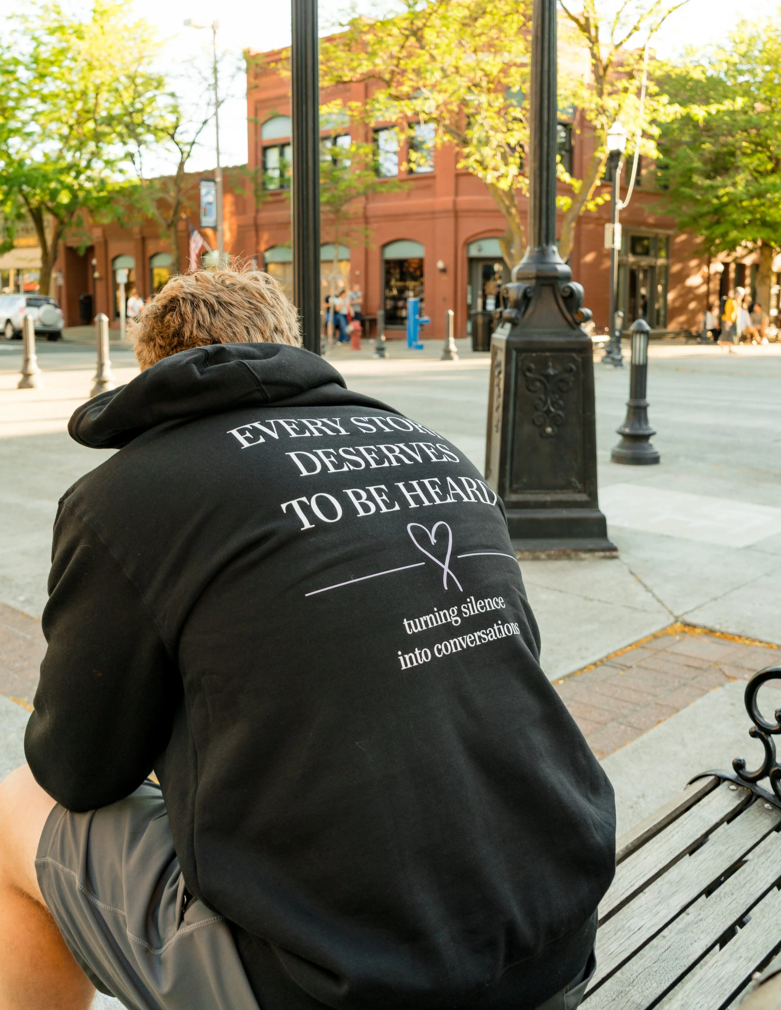 A person with blonde hair wearing a black hoodie with white text sitting on a park bench, in an urban setting with trees and a brick building in the background.