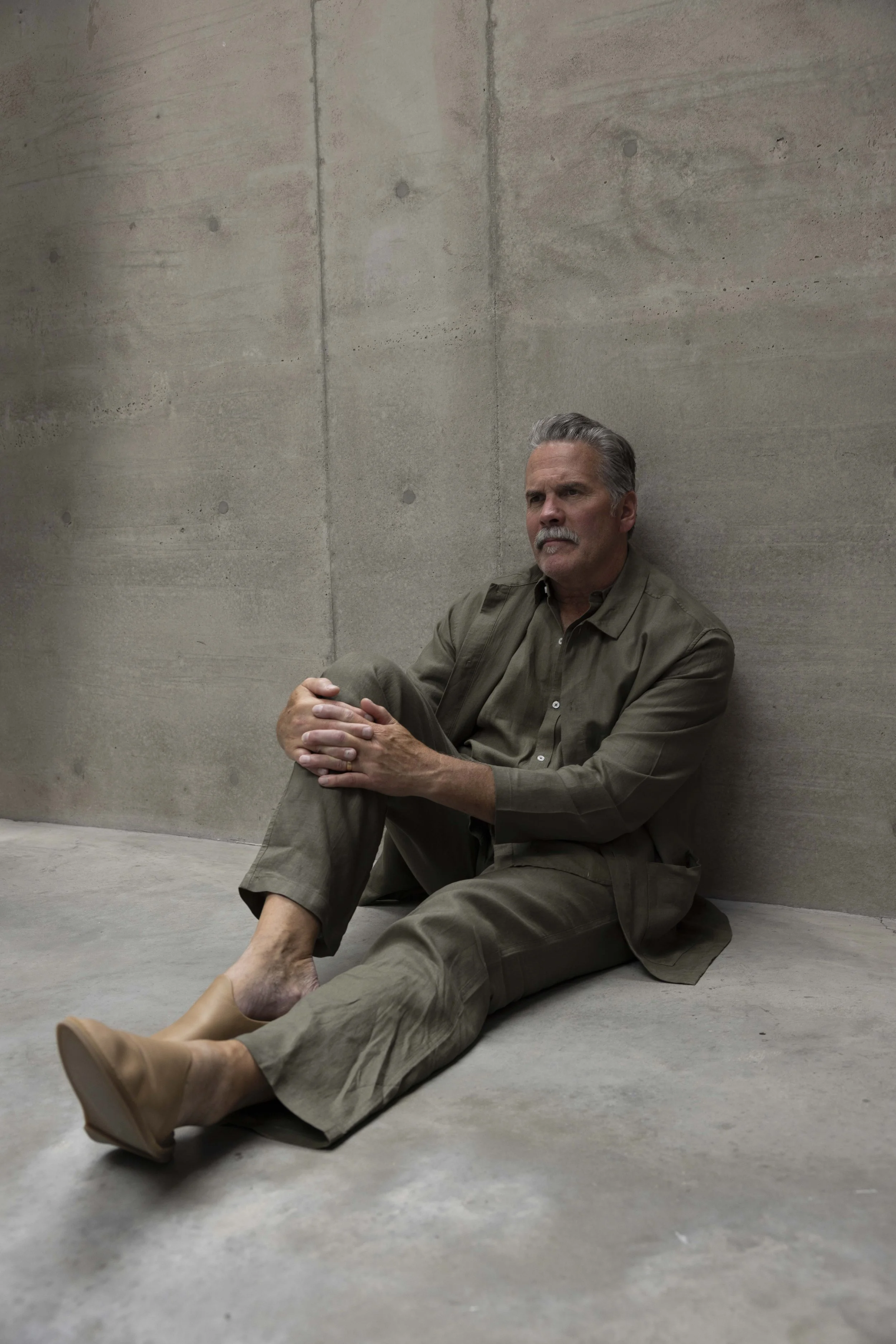 A middle-aged man with gray hair and a mustache sitting on the floor against a concrete wall, dressed in olive-green clothing and tan boots, looking thoughtfully to the side.