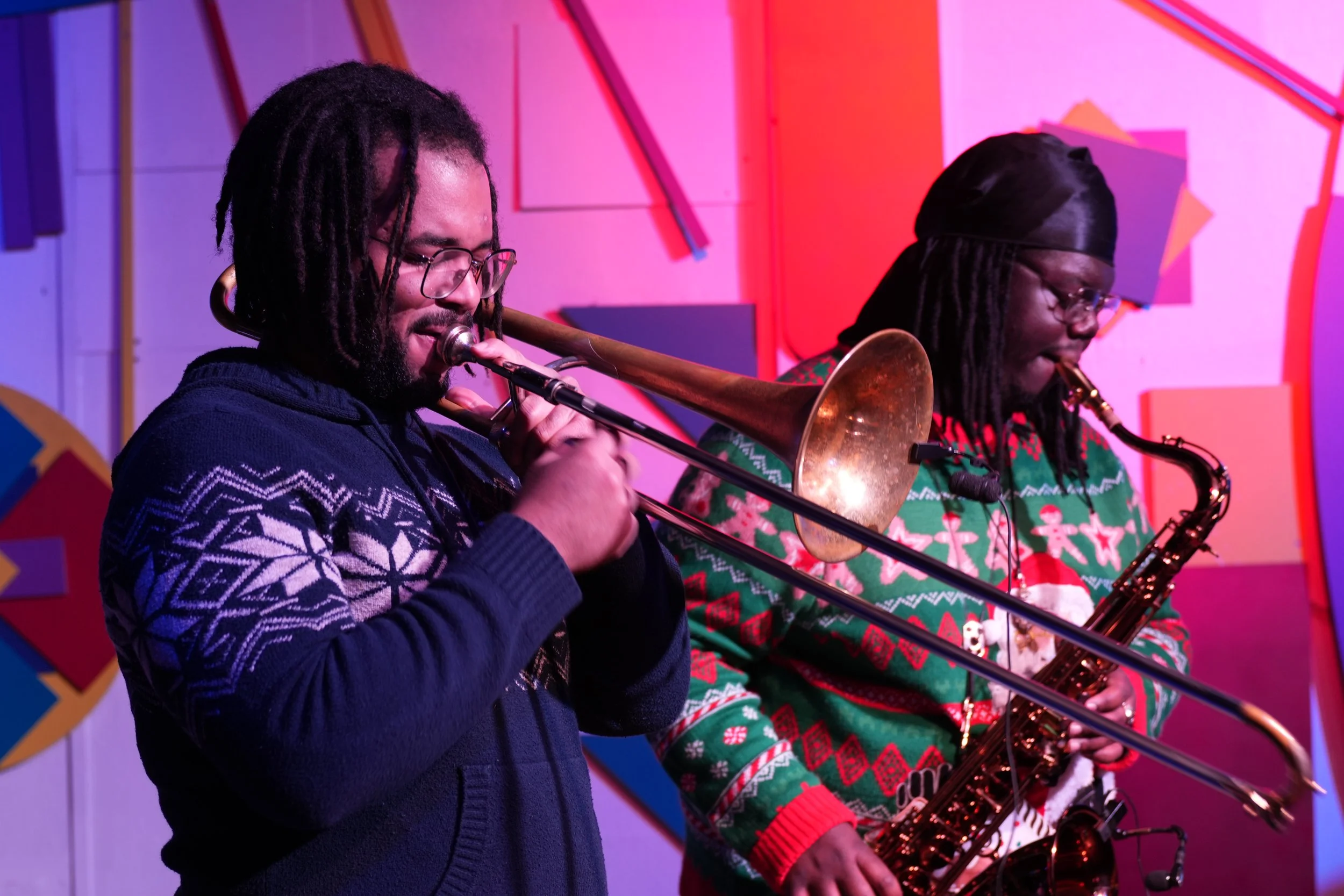 Two people playing musical instruments, one with a trombone and the other with a saxophone, in festive holiday sweaters, in front of a colorful geometric background.