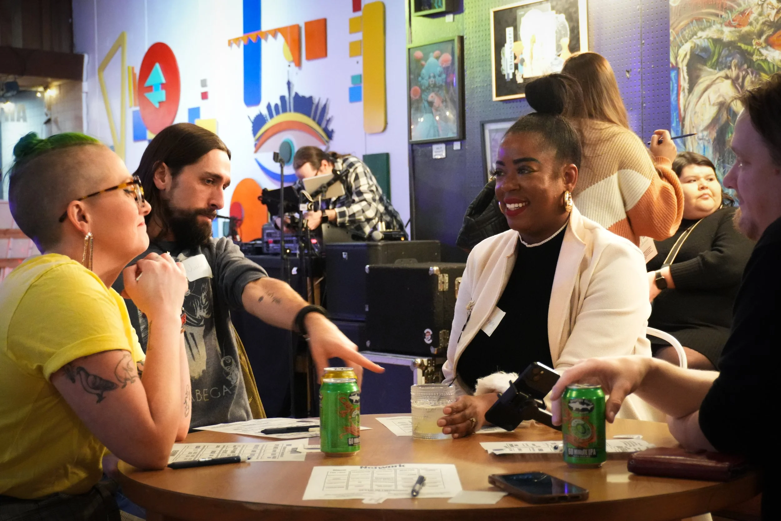 People sitting around a table at an event or gathering, engaging in conversation. The background features colorful artwork and walls, with cans and papers on the table.