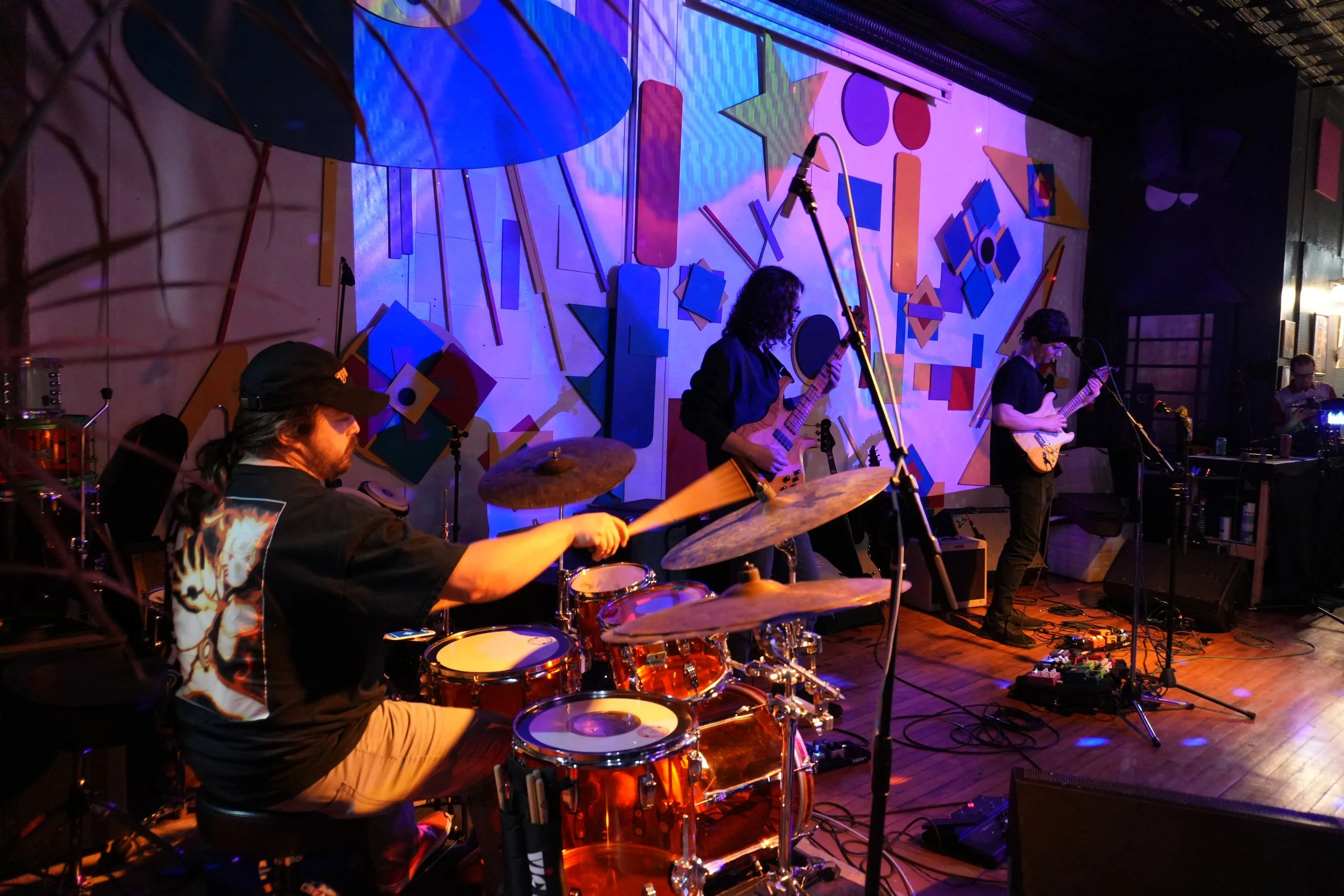 A live band performing on stage at The Rocki Button, with musicians playing drums, guitar, and keyboard, in a colorful, artistic setting with abstract geometric shapes on the wall.