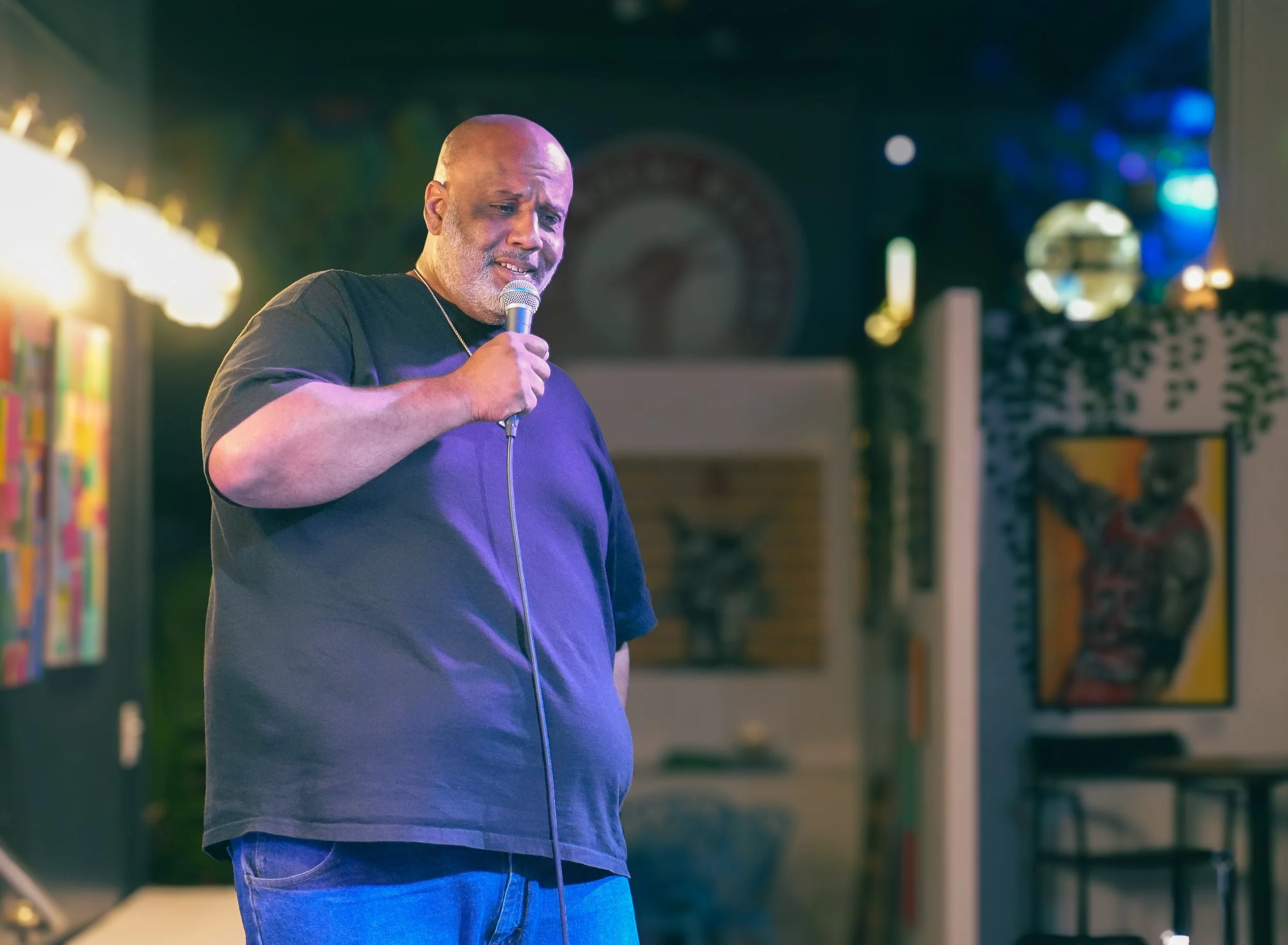 A middle-aged man holding a microphone and singing in a dimly lit indoor space. He is wearing a black t-shirt and blue jeans, with a serious expression on his face. The background includes colorful lights and what appears to be artwork or posters on the walls.