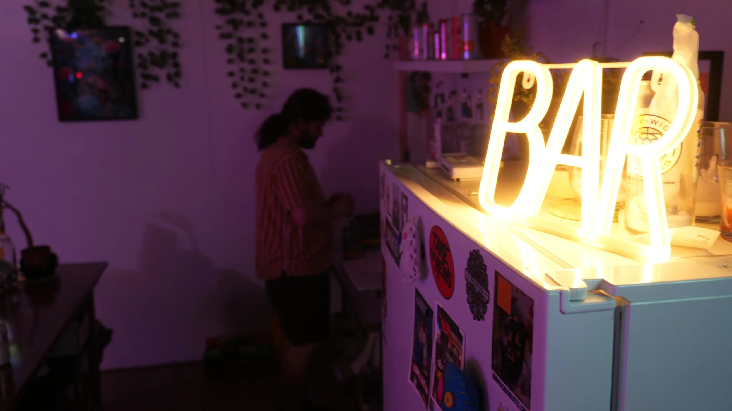 A person with dark hair wearing a red and white striped shirt stands in a room with purple lighting. There is a large illuminated sign that reads 'BAR' on a counter. The room has decorated walls with pictures and a bookshelf with colorful books.