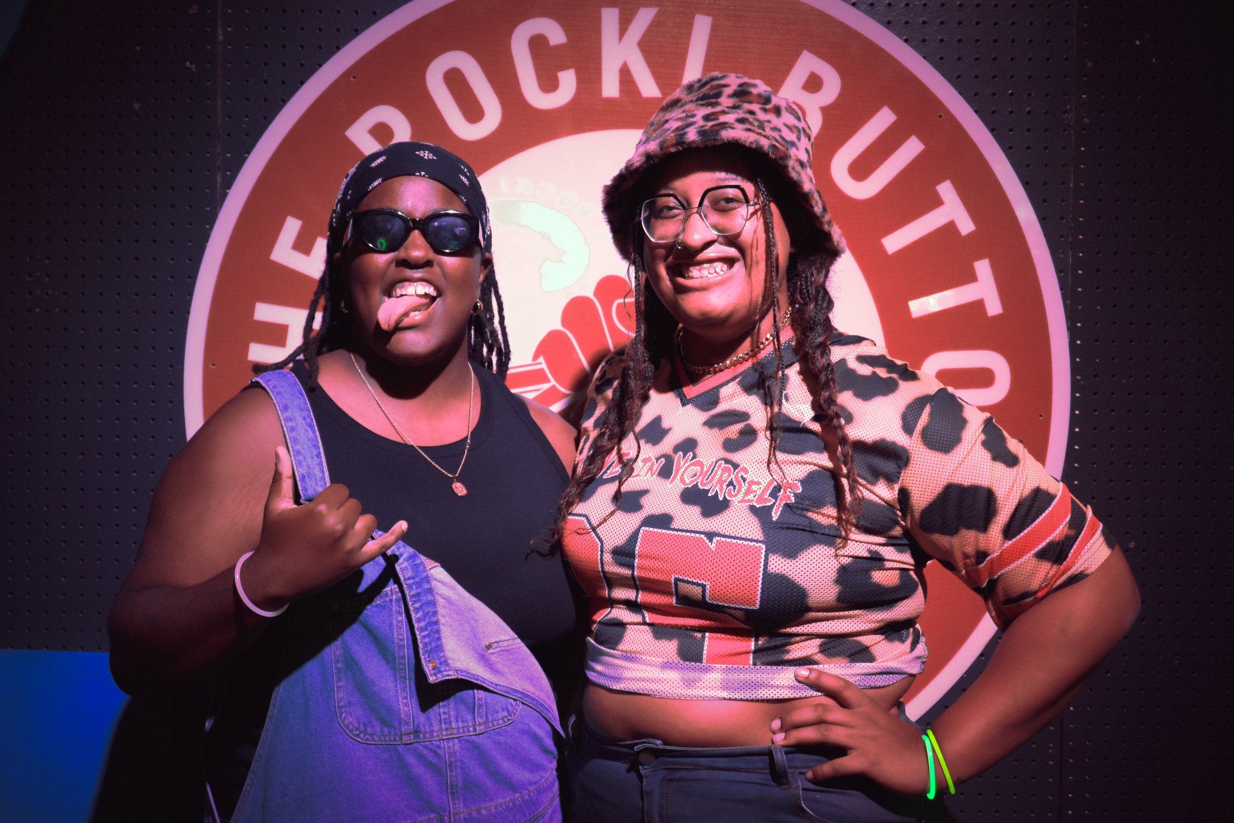 Two women smiling and posing for a photo in front of a circular sign that says "PUCK & BUSH TET". The woman on the left is wearing sunglasses, a black bandana, and a sleeveless top with denim overalls, sticking out her tongue and making a rock gestur