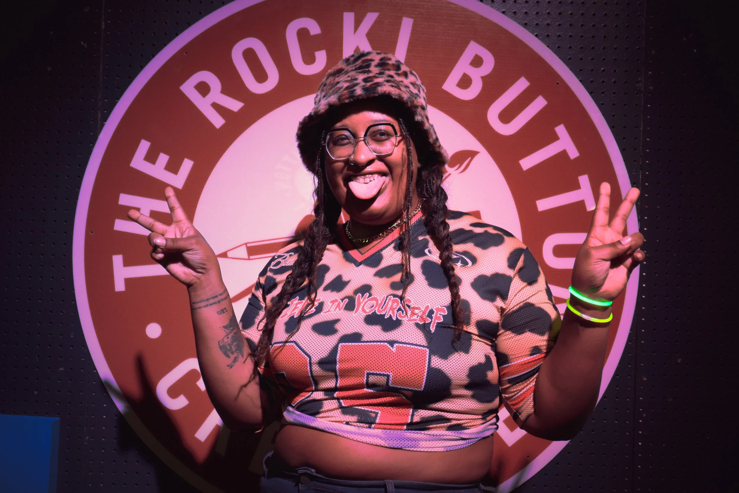 A person with long braided hair, glasses, and tattoos, wearing a leopard print hat and a sports jersey with the words 'Believe in Yourself,' making peace signs with both hands, standing in front of a circular sign that reads 'Hatch Rock'.