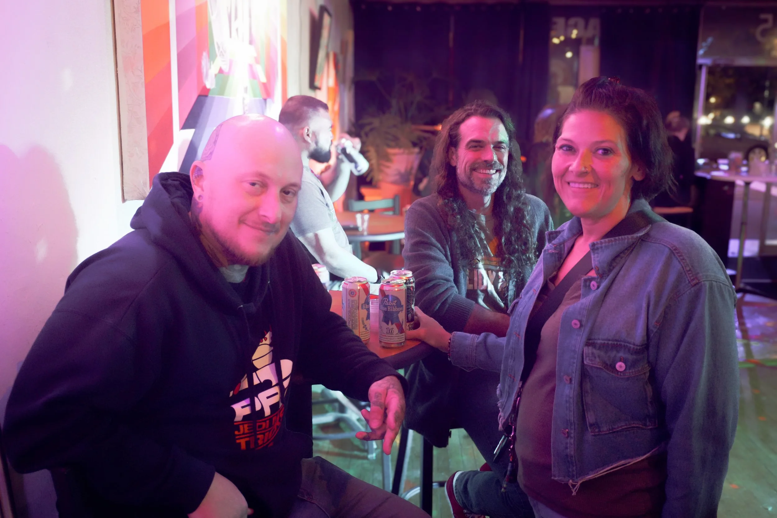 Four friends sitting at a table in a vibrant bar or nightclub, smiling and enjoying drinks, with colorful wall art and dim lighting in the background.