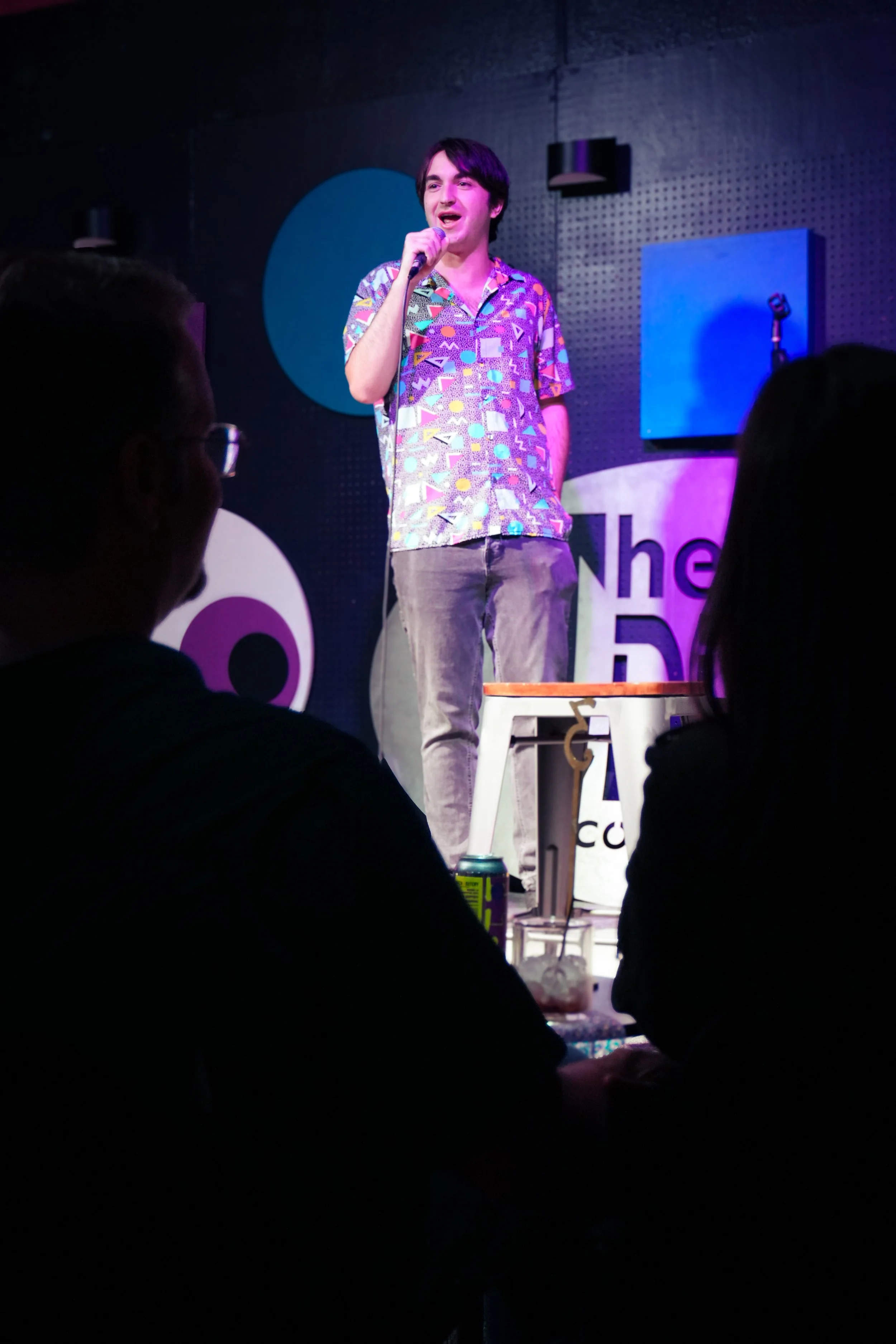 A young man with dark hair, wearing a colorful patterned shirt, stands on stage speaking into a microphone during a comedy show or stand-up performance. The background shows a black wall with purple and blue lighting, and there's an audience seated i