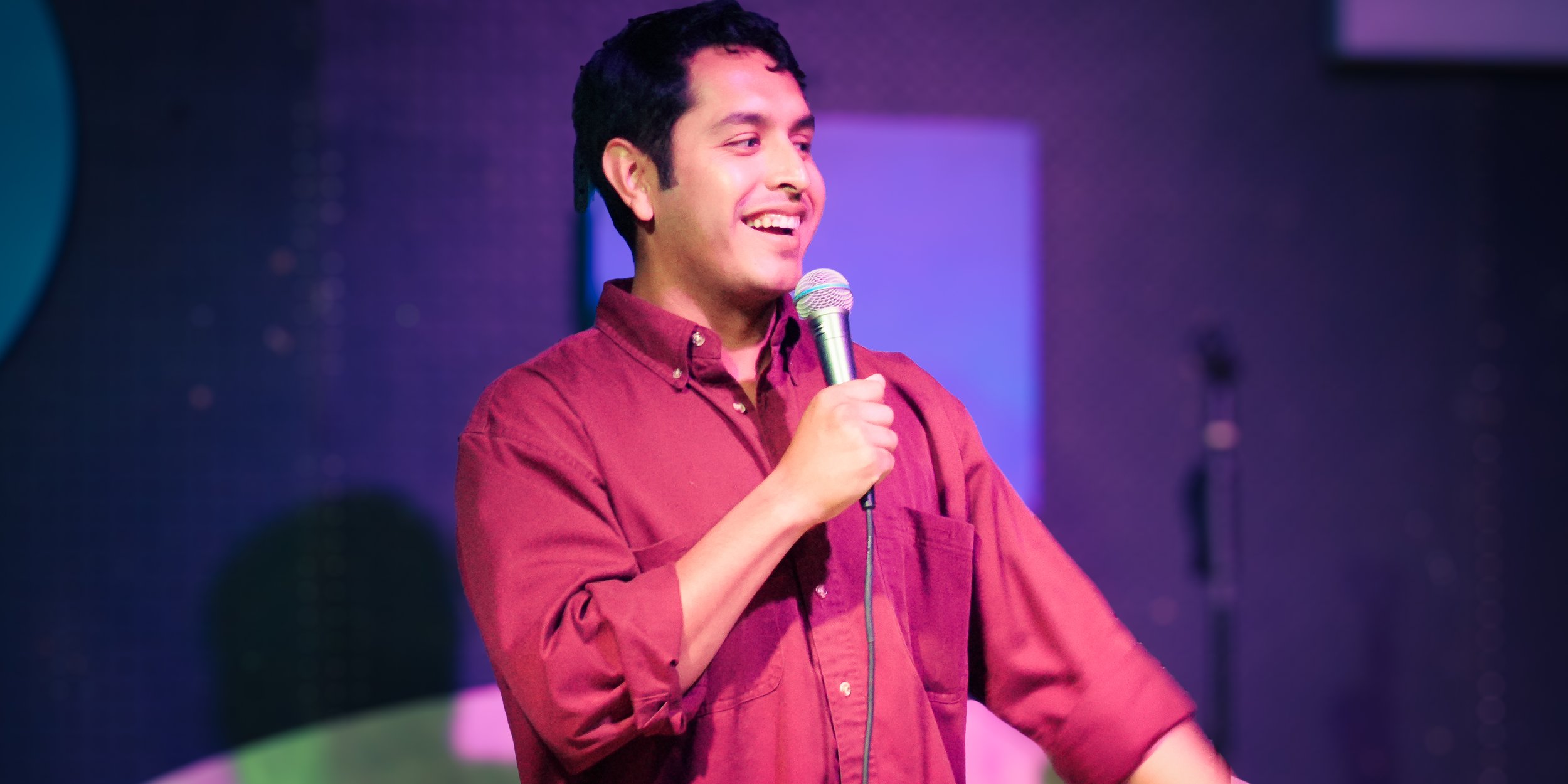 A man with short dark hair, wearing a red button-up shirt, is holding a microphone and smiling on stage with purple lighting in the background.