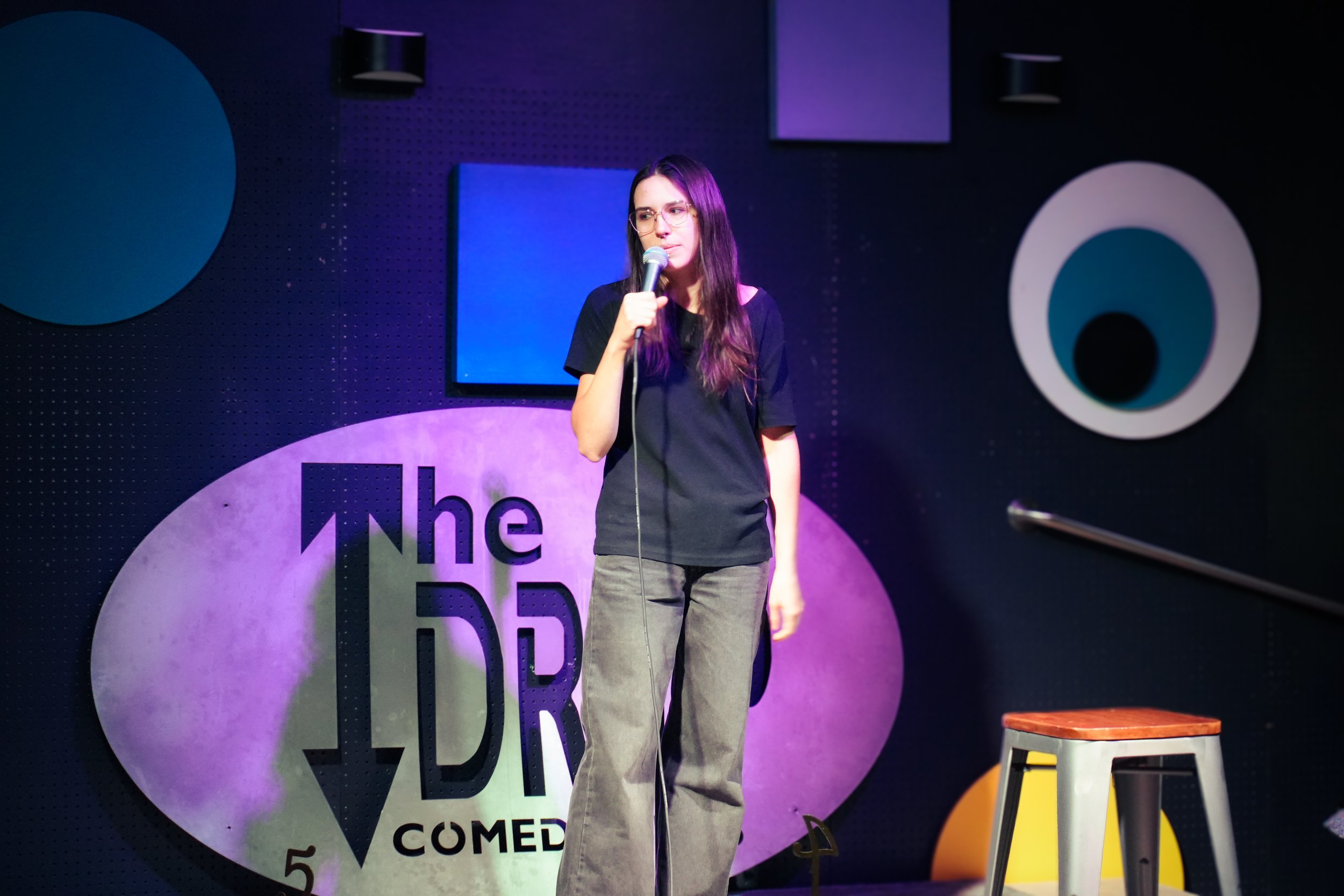 A woman with long dark hair, glasses, wearing a black t-shirt and gray pants, holding a microphone, standing on stage at a comedy show with a black background and colorful circular decorations.