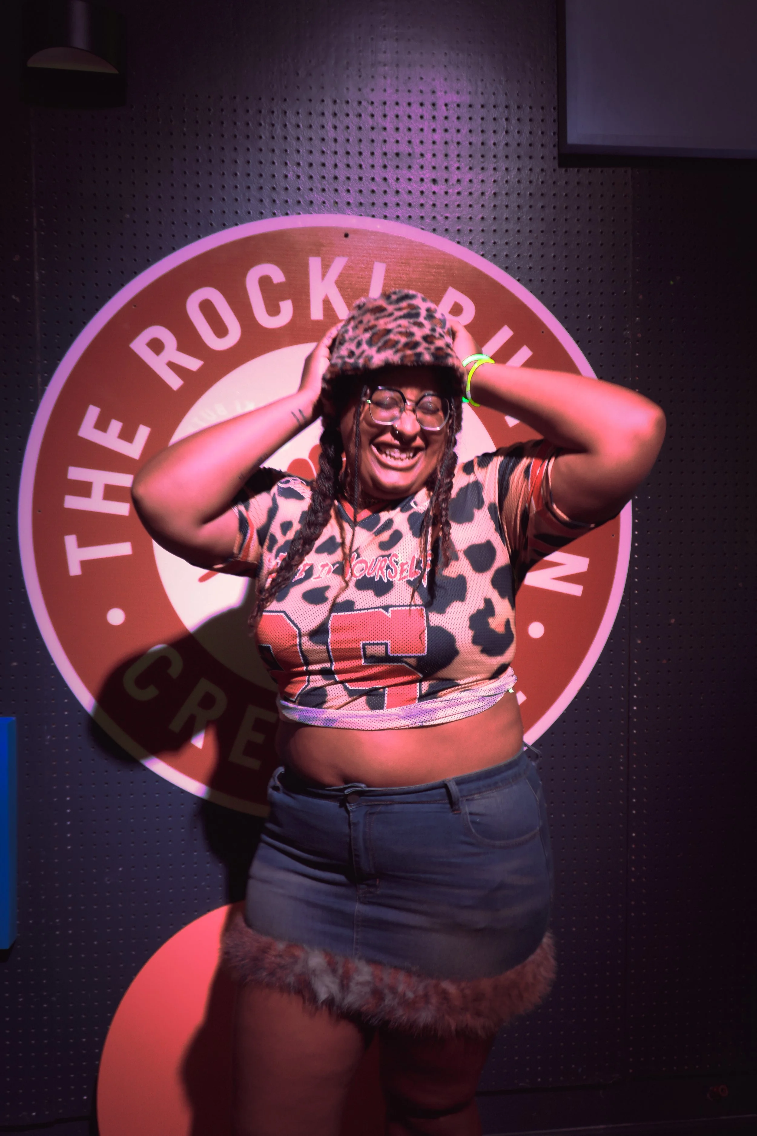 A person with glasses and braids wearing a leopard print hat, a leopard print top, and furry shorts, smiling and holding their head with both hands, standing in front of a backdrop with a circular logo that reads 'The Rock'.
