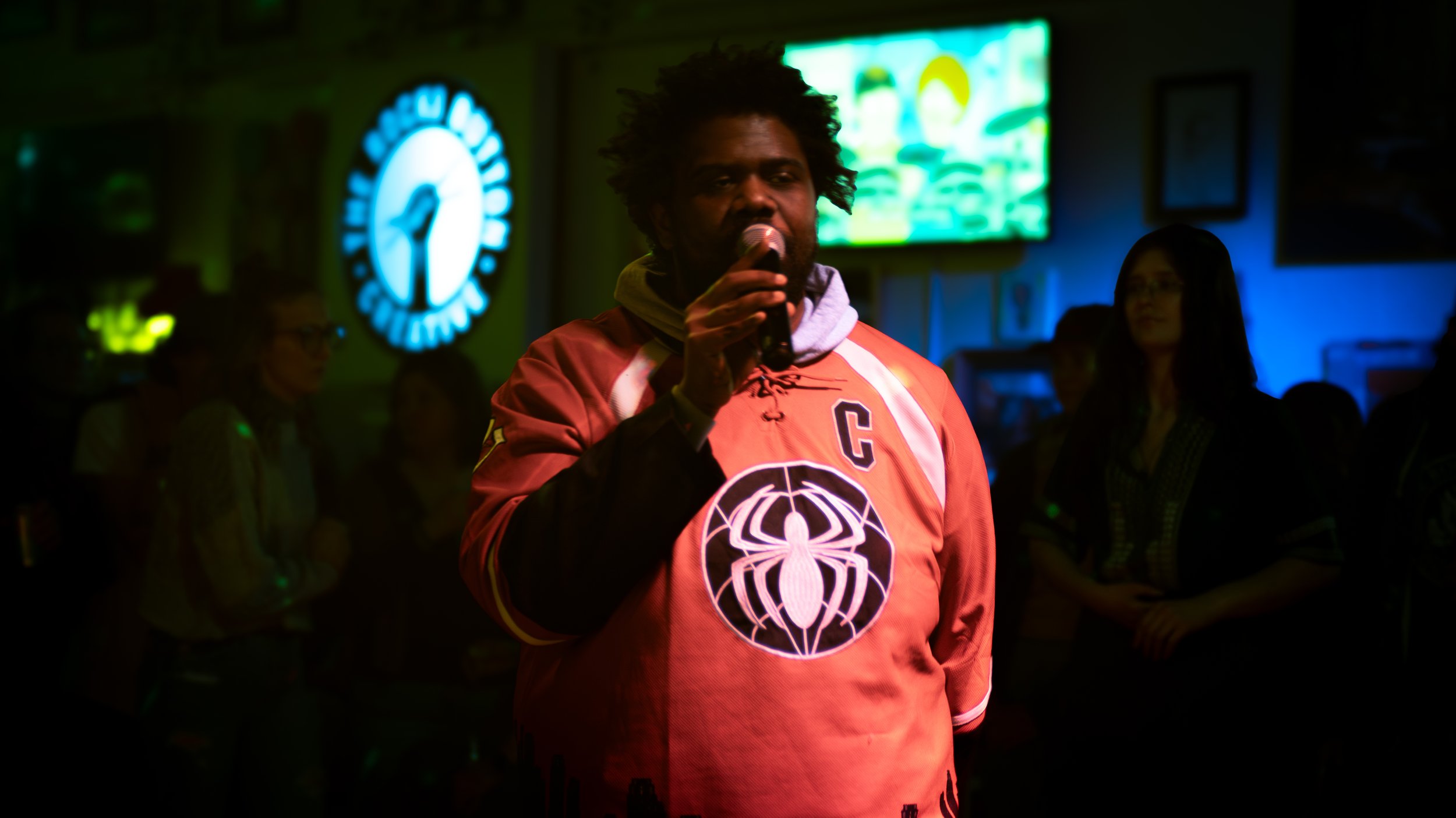 HEYZEUS holding a microphone, wearing a red hoodie with Spider-Man logo, standing in a dimly lit room with neon signs and a screen in the background.